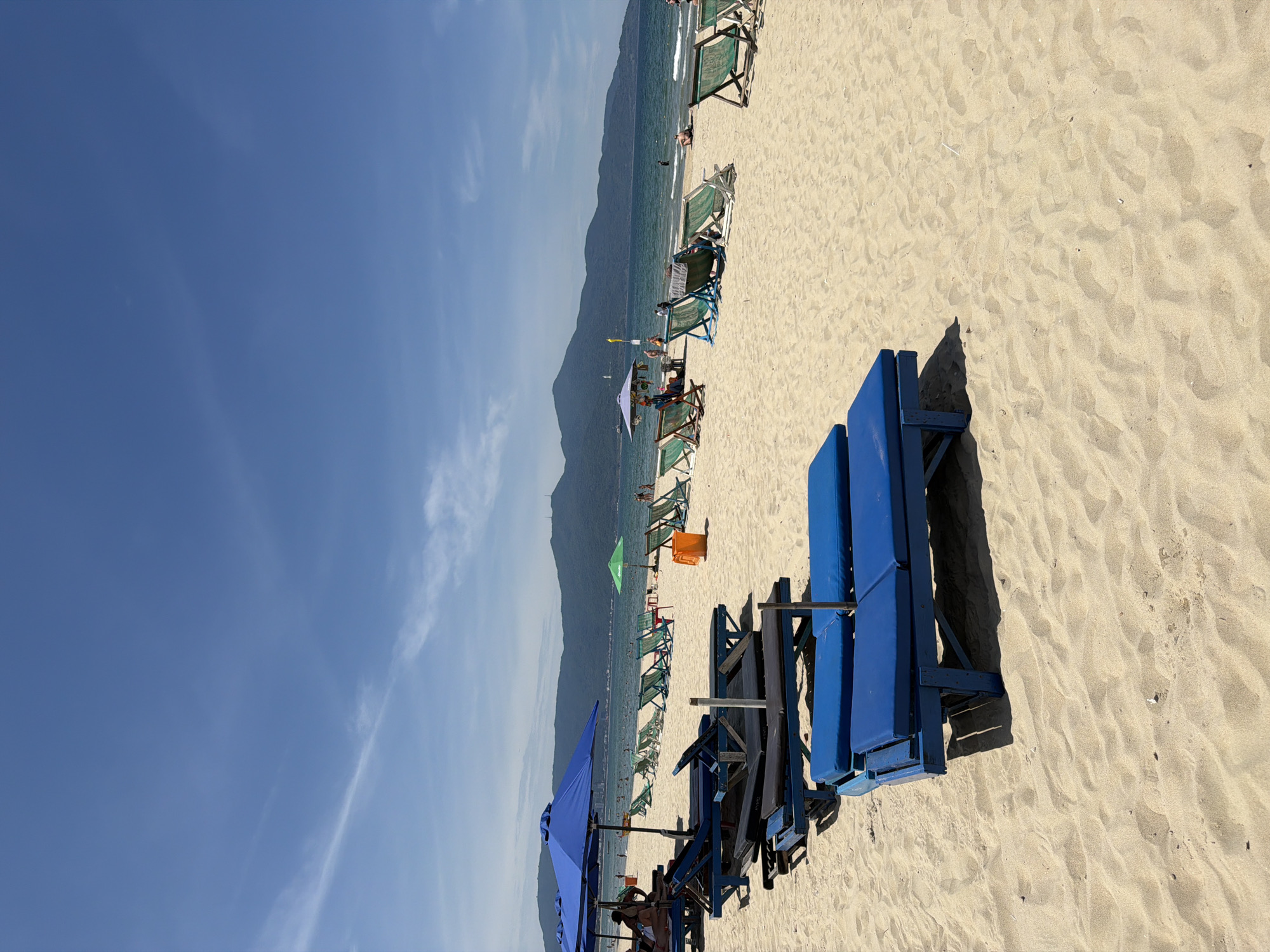 This is a sun-drenched beach scene in Da Nang, Vietnam, likely My Khe Beach, given the presence of the Marble Mountains in the background. The foreground is dominated by rows of empty blue lounge chairs, some with cushions, set on pale, fine sand. A large blue umbrella shades a portion of the sand in the mid-left. Further back, a line of green beach chairs and several more umbrellas are visible. People are scattered sparsely along the shoreline and in the shallow waters of the turquoise sea. The horizon is marked by a range of green, tree-covered mountains under a bright blue sky with thin, wispy clouds. An orange trash can stands near a green umbrella in the mid-ground. The overall impression is one of a quiet, pleasant day at the beach.