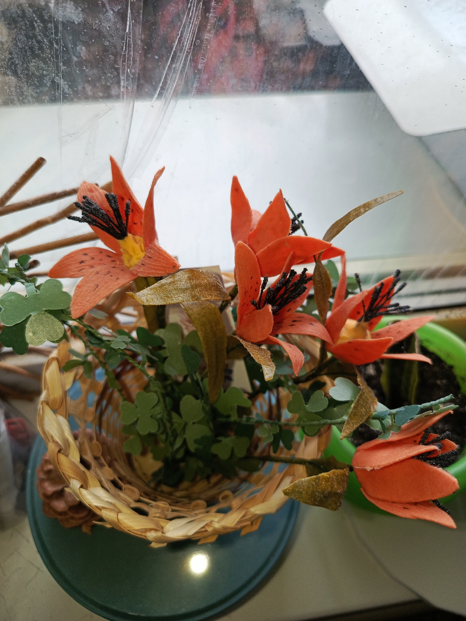 This is a close-up, high-angle, indoor shot of a decorative arrangement of artificial orange lilies with black stamen. The lilies are clustered in a woven, light brown basket, which sits on a teal-colored, circular plate. The basket contains green, clover-like artificial leaves and some dry, brown leaf-like accents. Behind the basket, part of a green planter with dark soil and a hint of a plant with striped leaves is visible. The background is out of focus, showing a window with some condensation or plastic sheeting on it. The lighting suggests it is daytime, possibly overcast, with some diffused light coming from the window. The scene is static and appears to be on a windowsill or a table near a window. There are no people or animals present. The context of Yakutsk, Russia is not visually evident in the image.