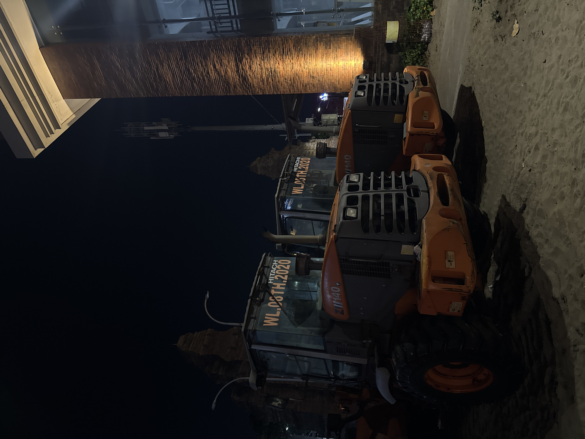 The image captures a nighttime scene in Kuta, Indonesia, featuring two orange and grey Hitachi wheel loaders parked on a sandy surface, likely a construction site or an area undergoing development. The loaders are positioned side-by-side, with the one on the left slightly closer to the foreground. Both machines have visible markings, including "HITACHI," "WL.08TH.2020," and "ZI//140" on their exteriors. The cab windows of the left loader are illuminated from within, suggesting the presence of interior lighting.
Behind the loaders, a tall, partially constructed brick structure with ornate, temple-like elements is visible against the dark night sky. To the right of the loaders, a modern building with a brick facade is illuminated by warm, golden lights, highlighting the textured surface of the bricks. A large window or glass facade is also present on this building, revealing a dark interior. A cell tower stands prominently in the background, to the left of the modern building. The ground is covered in sand, with tire tracks from the heavy machinery clearly imprinted. The overall atmosphere is industrial and somewhat desolate due to the nighttime setting and the presence of heavy construction equipment.