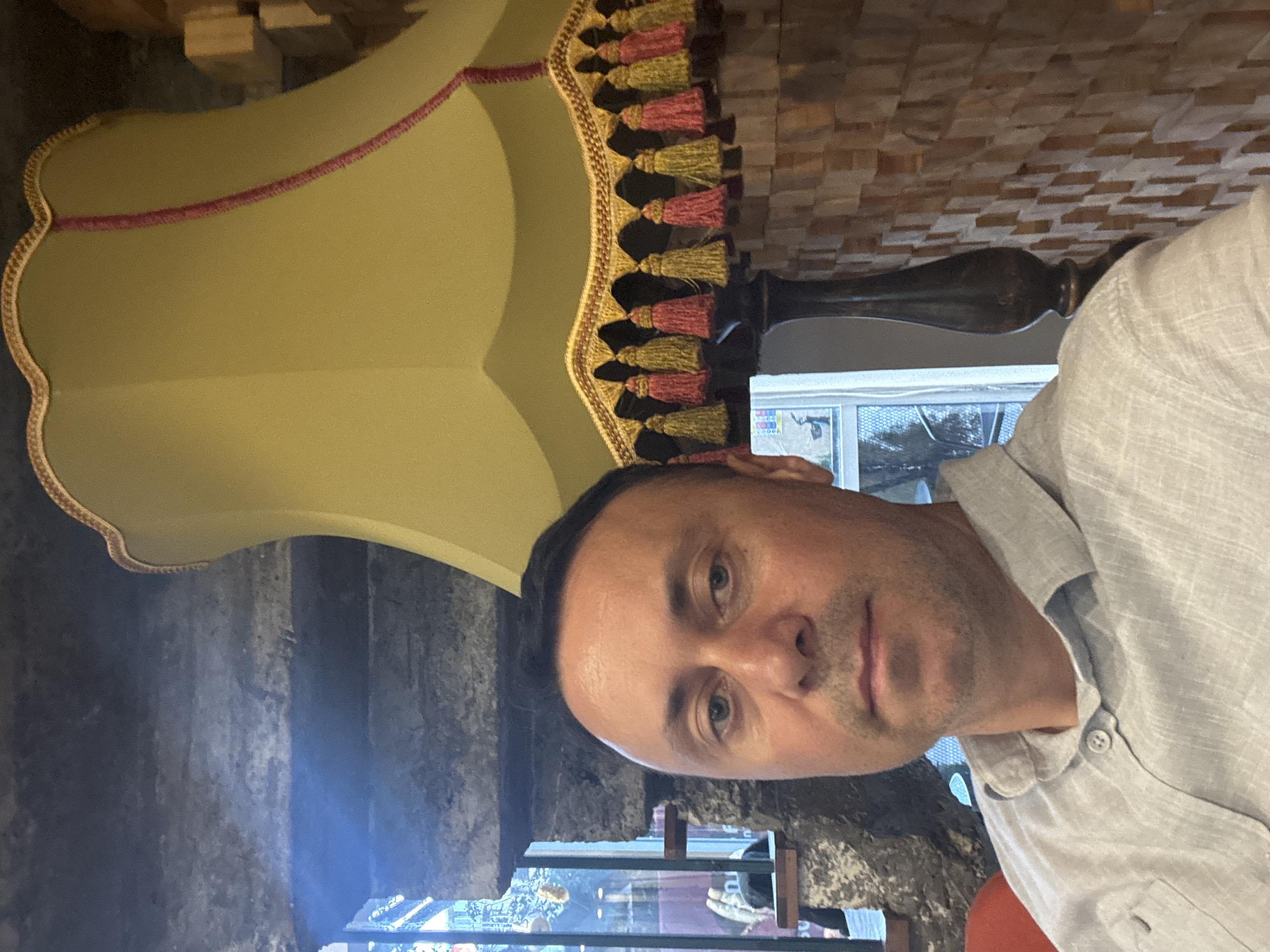 The image shows a man with short, dark hair and light skin, wearing a light-colored collared shirt. He is seated in what appears to be an indoor setting with a modern, industrial aesthetic. Behind him, a large, pale yellow lampshade with decorative tassels and trim is visible. To his left, a stone structure and a window can be seen, suggesting a possible location near a street or outdoor area. The background on the right features a textured wall made of stacked wooden blocks. The overall lighting is dim, with the primary light source appearing to be natural light coming from the window. The man is looking directly at the camera. The scene is not identifiable as being in Tbilisi, Georgia.