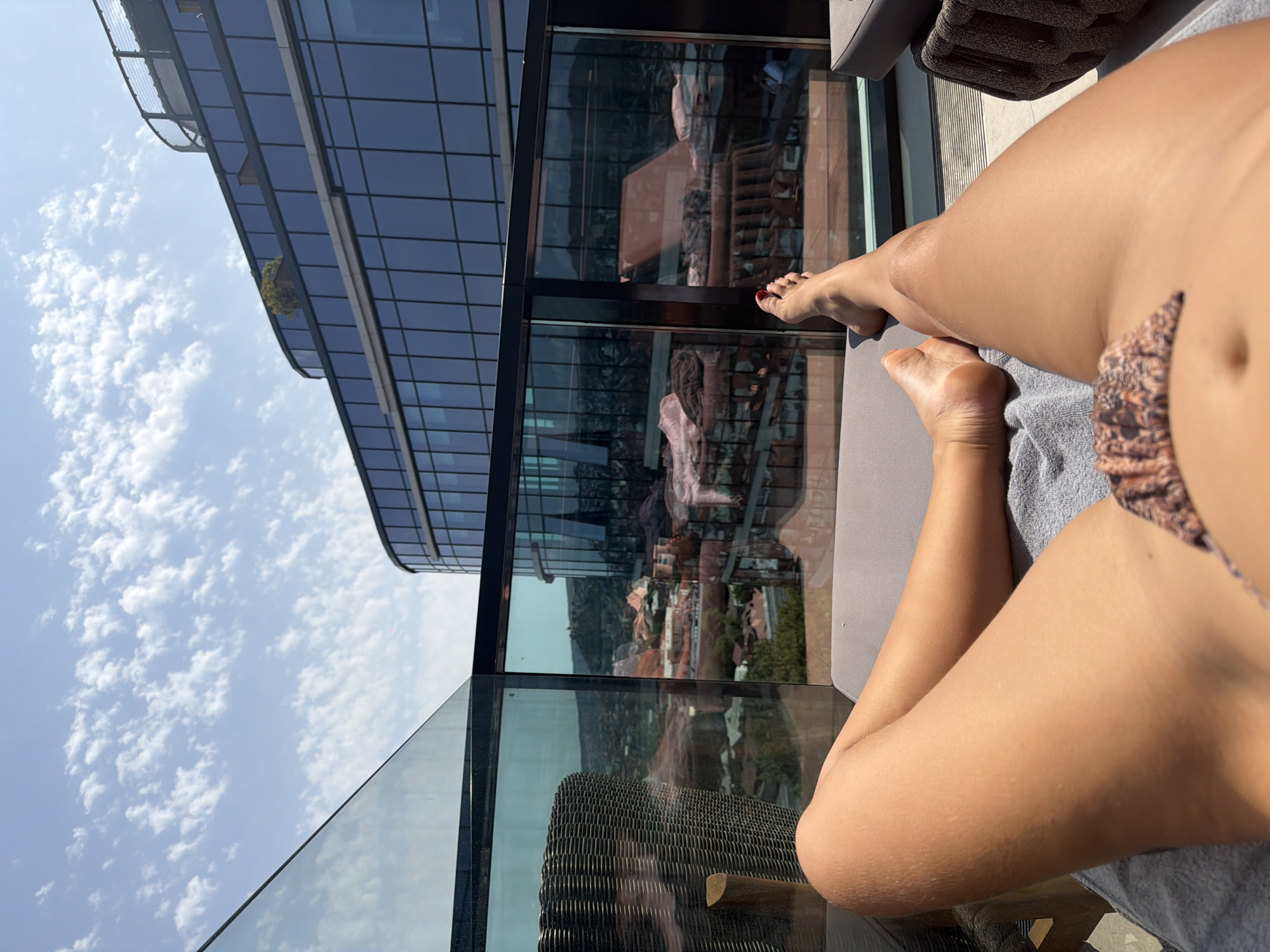 The image shows a first-person perspective of a person relaxing on a lounge chair on a rooftop balcony in Tbilisi, Georgia. The person's legs and lower torso are visible in the foreground, wearing a patterned bikini bottom. The background features a modern building with large glass windows, reflecting the sky and surrounding cityscape. The city's architecture and hilly terrain are visible beyond the balcony, under a bright, cloudy sky.