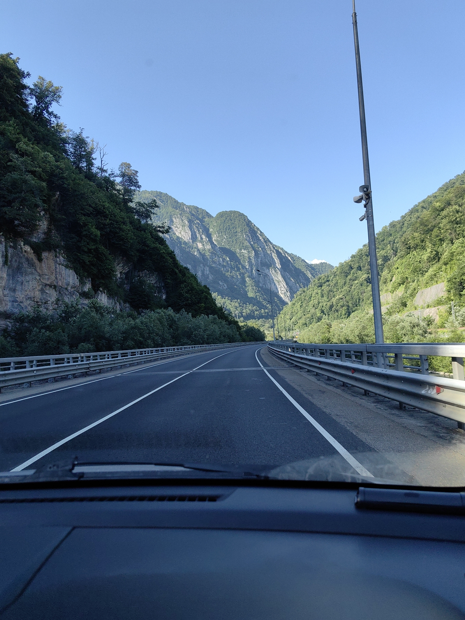 The image captures a view from inside a vehicle driving on a paved road through a mountainous, forested region. The road has two lanes in the direction of travel, with white lane markings. A metal guardrail runs along the right side of the road. On the left, a steep, rocky cliff face is covered with dense green foliage. On the right, a similar steep, forested mountainside rises. A tall pole with surveillance equipment is visible on the right side of the road. The sky is clear and blue. The scene appears to be an outdoor landscape with natural scenery and infrastructure for transportation. The specific location in the city of Monastyr, Russia is not identifiable from the visual information in the image.