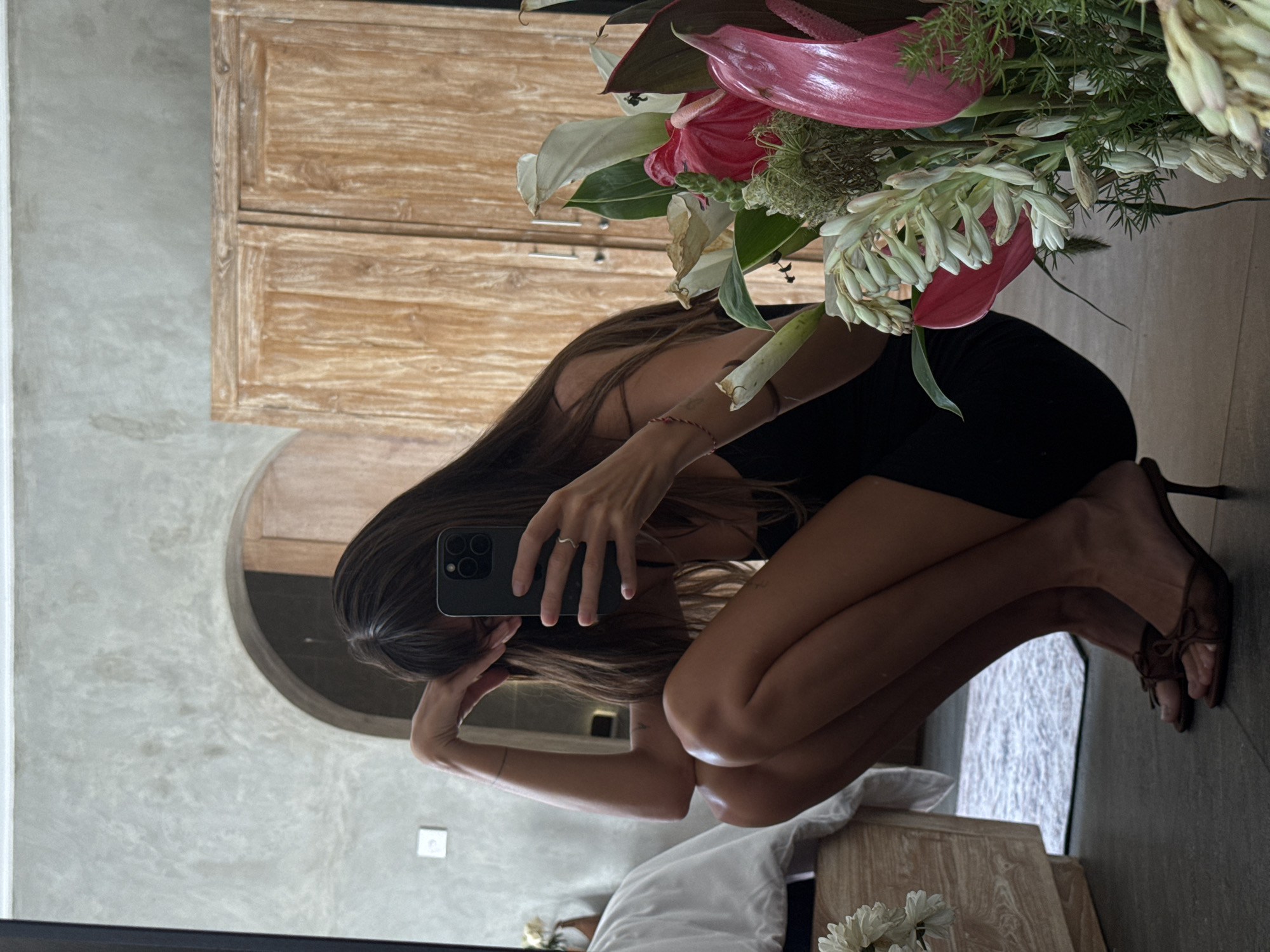 A woman crouches in front of a mirror taking a selfie with her phone, obscuring her face. She wears a black dress and brown high-heeled sandals. A bouquet of flowers partially blocks the view of her upper body. The reflection shows a bathroom interior with a wooden cabinet and arched doorway. The scene is located in North Kuta, Indonesia.