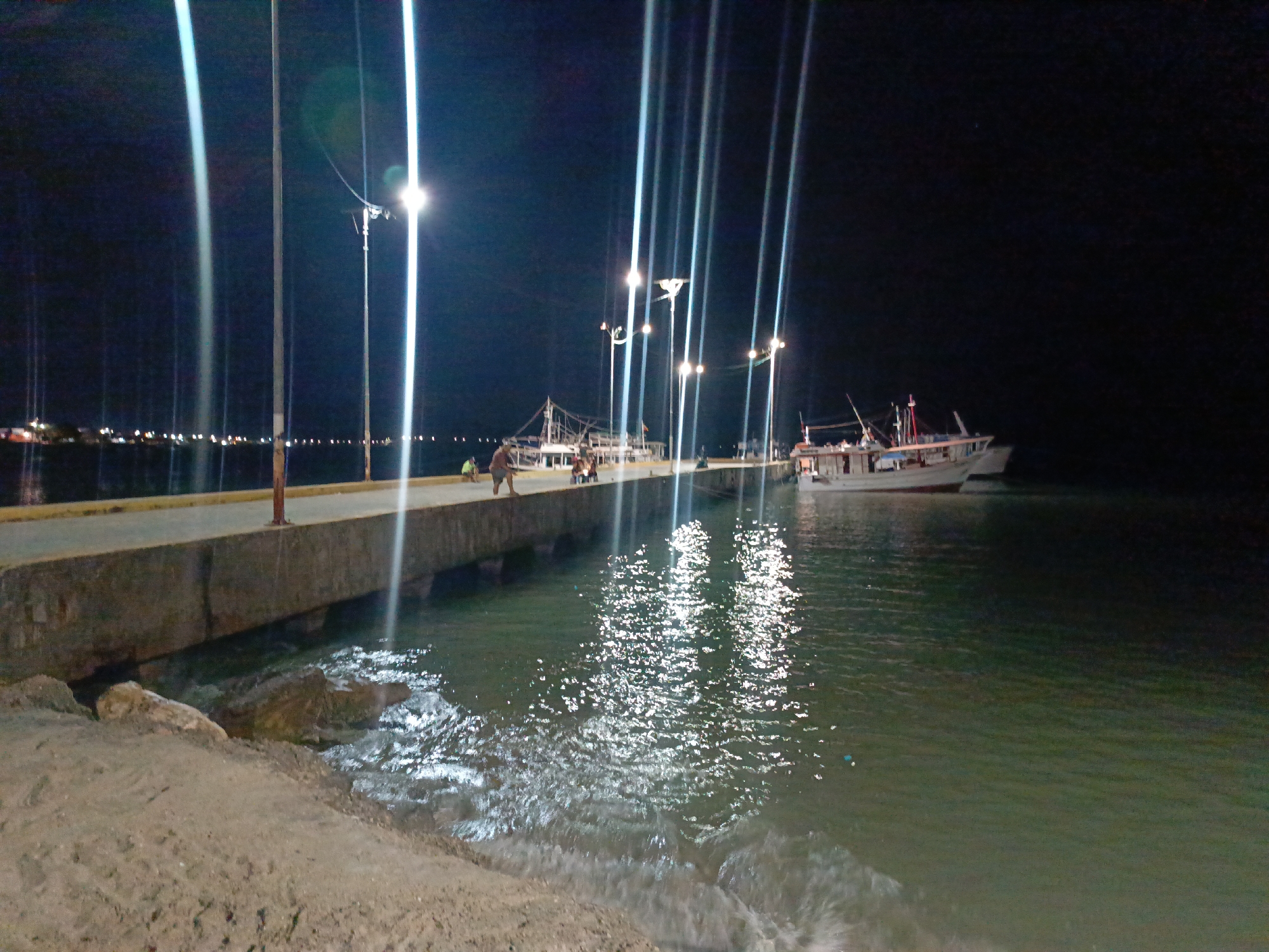 The image depicts a nighttime scene at the harbor of Juan Griego, Venezuela. A concrete pier juts into the water, illuminated by a series of tall lampposts. The lights cast bright reflections on the water's surface. Several people are visible on the pier. A fishing boat is docked alongside the pier. The foreground shows a sandy beach with gentle waves lapping the shore. The night sky is dark, with faint lights visible in the distance across the water.