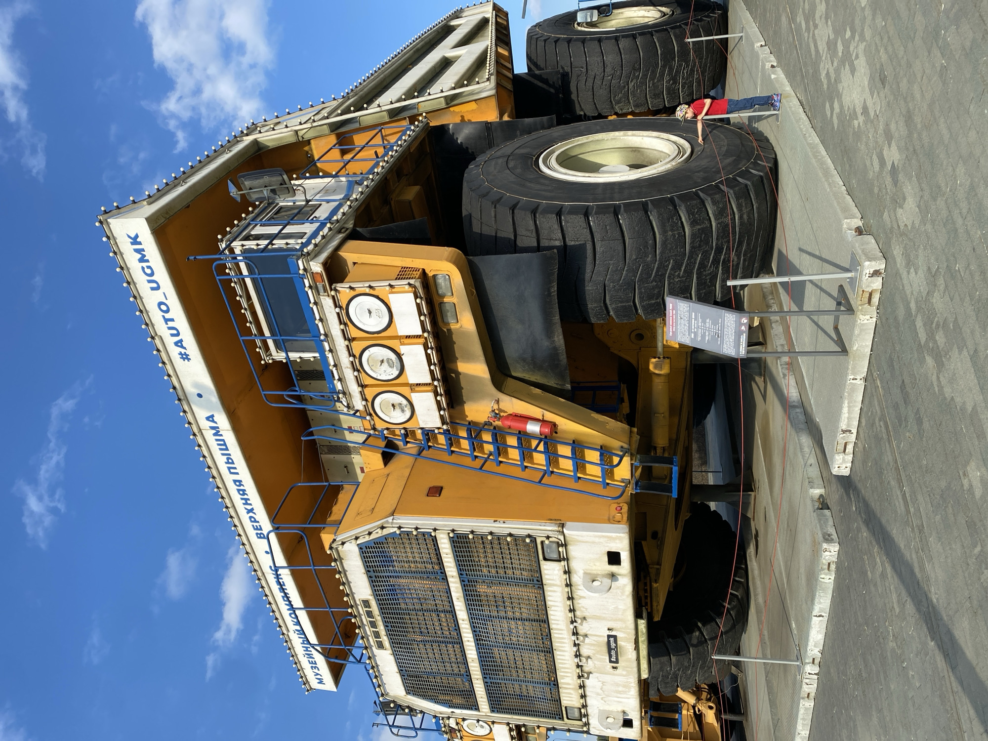 This is a photograph taken outdoors under a blue sky with scattered clouds. The main subject is a very large, yellow mining truck, likely a BelAZ, displayed as part of a museum complex. The truck is predominantly yellow with white and blue accents. It has massive black tires. Blue railings and stairs are visible on the side of the truck, leading up to the cab. A red fire extinguisher is attached to the stairs. The words "МУЗЕЙНЫЙ КОМПЛЕКС" and "ВЕРХНЯЯ ПЫШМА" (Museum Complex Verkhnyaya Pyshma) are written in blue text along the top edge of the truck. A hashtag "#AUTO_UGMK" is also visible in blue text.
In the foreground, a small child wearing a red shirt, blue jeans, and a patterned cap stands near the base of the truck, interacting with a display. The child is on a raised platform that is roped off with red cord. A metal information board is also present on the platform. The ground is paved with gray cobblestones. The overall scene suggests an outdoor exhibition or museum in Verkhnyaya Pyshma, Russia, showcasing large mining equipment.