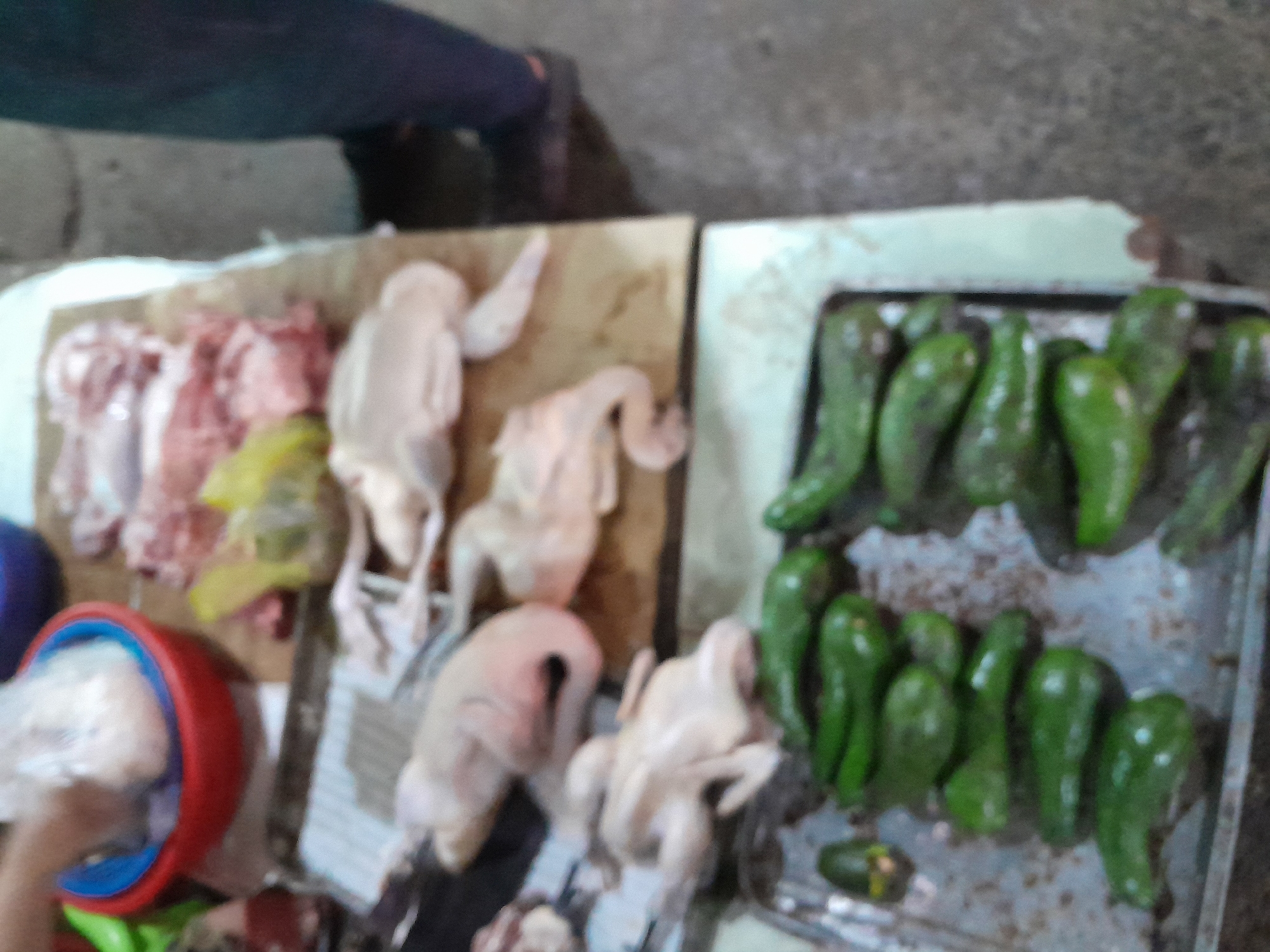 This is a medium shot, taken from a slightly elevated angle, of a market stall. In the foreground, a metal tray is filled with several rows of bright green, elongated vegetables, possibly chilies or small avocados. Behind the tray, on a wooden surface, are several raw, whole chickens laid out. Further back, there are cuts of raw red meat, also on the wooden surface. To the left, a red bowl with a blue rim contains what appears to be more meat wrapped in plastic. A person's hand, holding a plastic-wrapped package, is visible at the top left of the frame. The background is out of focus but suggests a market environment with a textured floor and a wall. The lighting is natural, casting soft shadows. The overall scene depicts a food market, likely in Mũi Né, Vietnam, showcasing fresh produce and raw meats for sale.