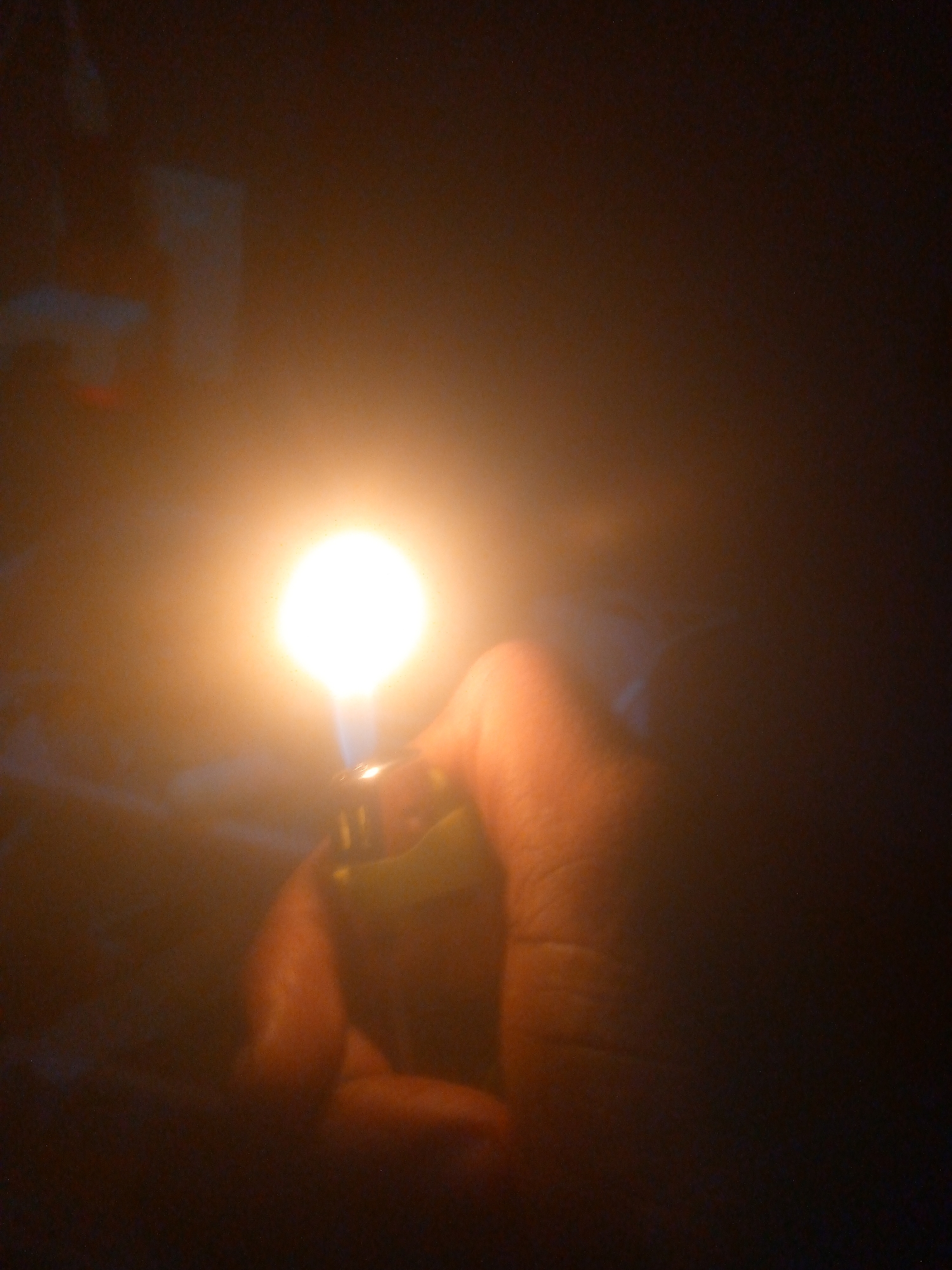 The image captures a close-up of a human hand holding a lit lighter in a profoundly dark environment. The bright, intense orange-yellow flame emanating from the lighter serves as the primary light source, illuminating the fingers and palm of the hand. The lighter itself appears to have a yellowish-green body.
The scene is dominated by shadow and warmth from the flame. The background is largely indistinguishable, appearing as deep shades of brown and black with faint, blurry textures or shapes visible in the upper left, hinting at unseen objects or an indoor setting. There are no discernible location cues, but the pervasive darkness suggests either nighttime or a completely unlit interior space.
The activity taking place is the act of igniting and holding a lighter, providing a singular point of intense light. The atmosphere is stark and intimate due to the extreme contrast between the bright flame and the enveloping darkness. There is no visible text in the image.