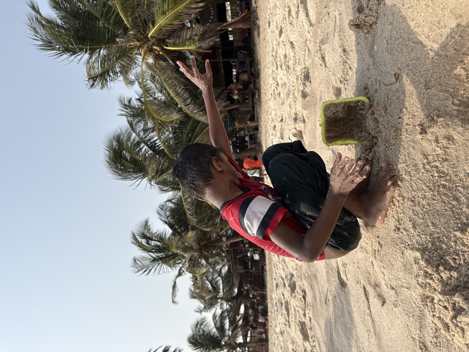 The scene depicts a young male child, likely pre-teen, crouching on a sandy beach under a clear, bright sky in Mopo Onijebu, Nigeria. He wears a red short-sleeved t-shirt with white and dark blue stripes on the sleeves, and dark trousers. His feet are bare, and sand adheres to his hands and arms. The child's head is tilted downward and to his left, while his right arm is extended straight out, fingers splayed and covered in sand. To his right, a light green plastic bucket, partially filled with sand, stands upright on the beach. In the midground, blurred figures of individuals are visible, some near rustic structures with thatched or tarp roofs. The background features numerous tall palm trees with green fronds, indicating a tropical coastal environment.