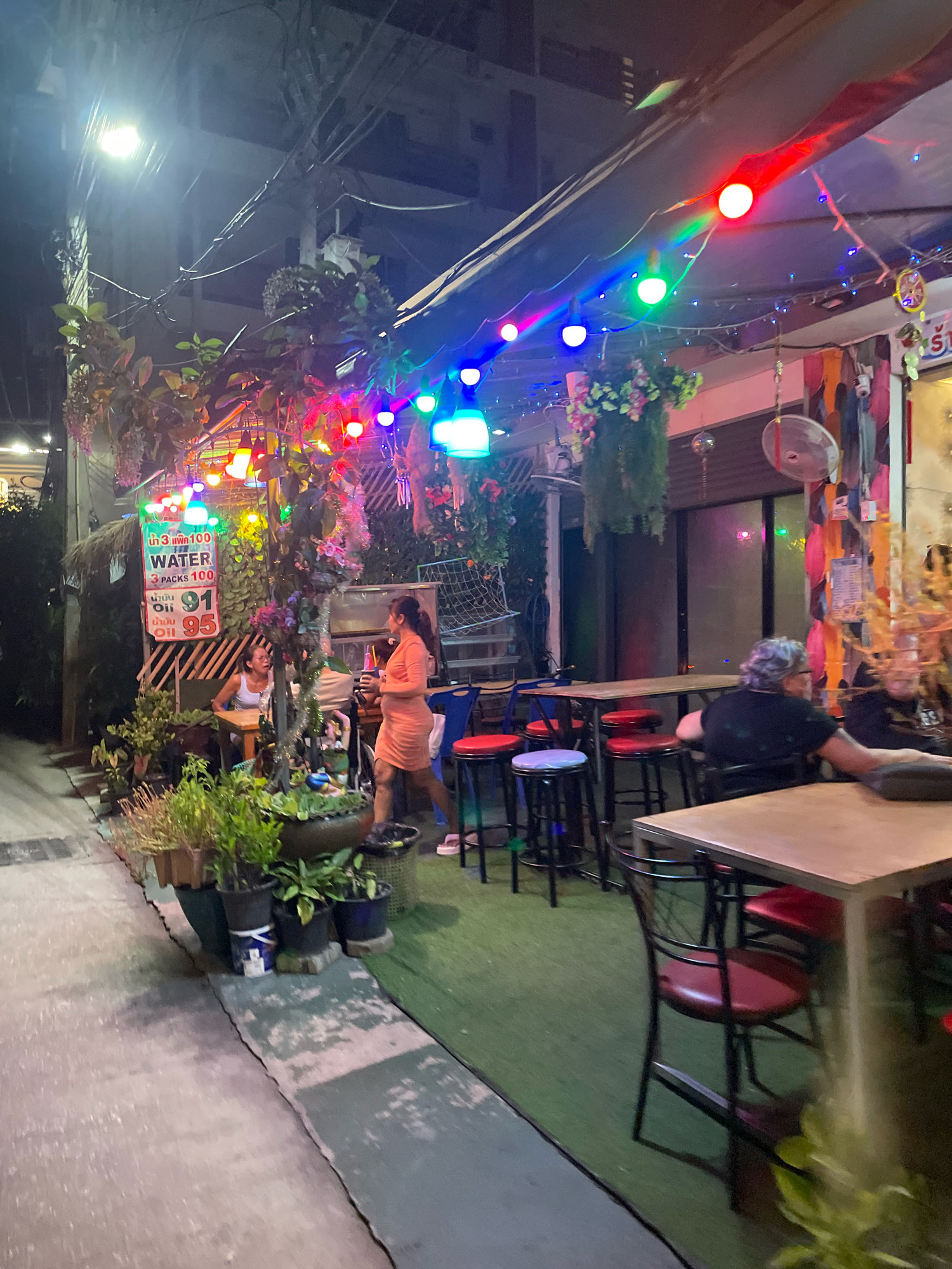 This image depicts a street-level view of a bar or restaurant in Pattaya City, Thailand, at night. Colorful string lights are draped along the awning and facade, illuminating the outdoor seating area. Several tables with chairs and stools are visible, some occupied by patrons. In the foreground, a woman in an orange dress stands holding a drink, facing away from the camera. To her left, another woman sits at a table with plants surrounding her. The street surface appears to be concrete, with a section of green artificial turf laid out in front of the establishment. The scene is dimly lit by the string lights and ambient city lighting, creating a vibrant and lively atmosphere.