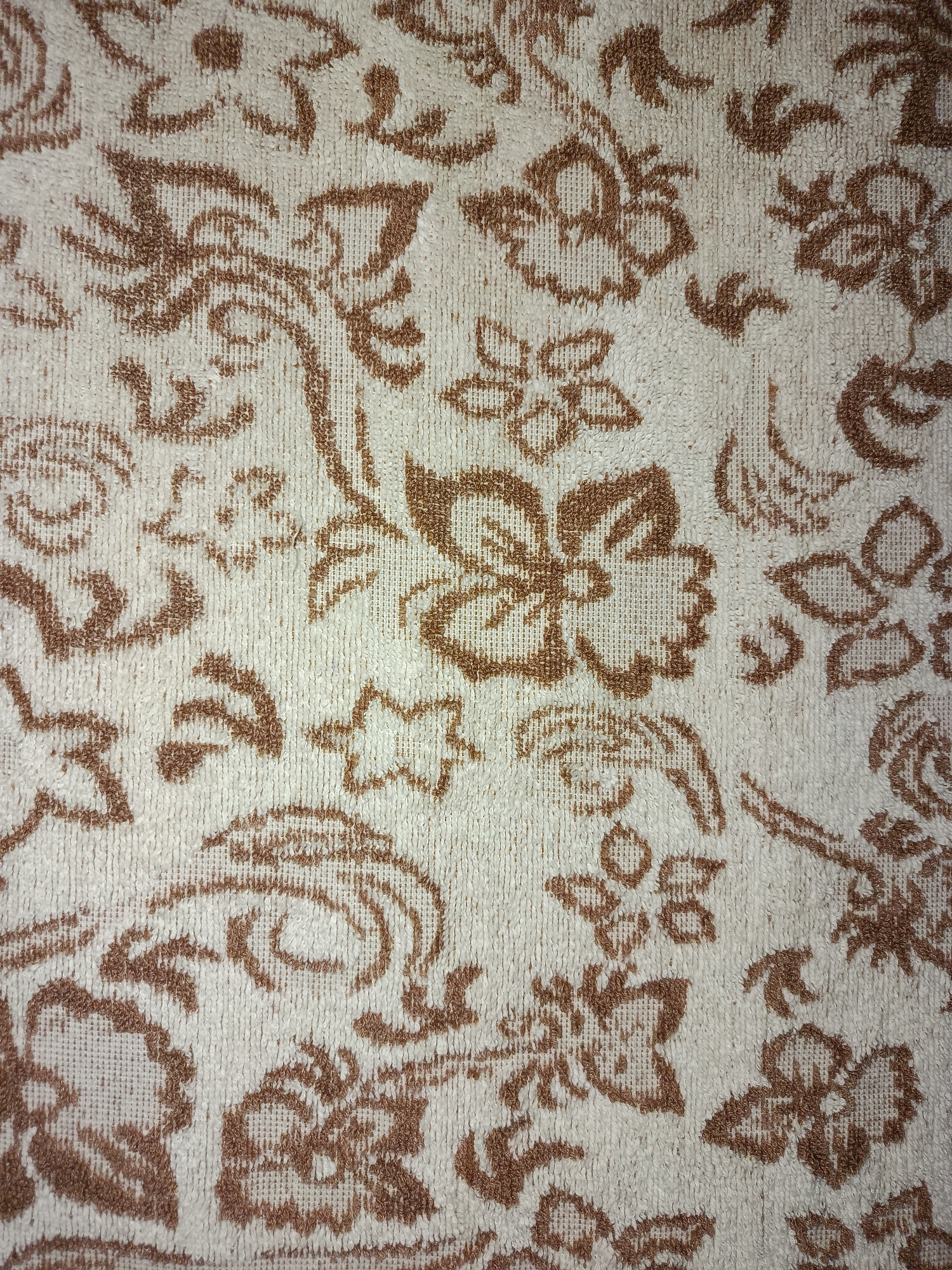This is a close-up, overhead shot of a fabric with a floral pattern. The background is a light beige, and the pattern is a medium brown, with motifs that resemble stylized flowers and vines. The fabric appears to be textured, possibly terrycloth, with visible loops. The lighting is even, suggesting it was taken indoors or in consistent light conditions. No people, specific objects, or scenery are depicted, only the texture and pattern of the textile. There is no visible text. The location context of Tashkent, Uzbekistan, provides no visual cues within the image itself. The image focuses solely on the decorative pattern of the fabric.