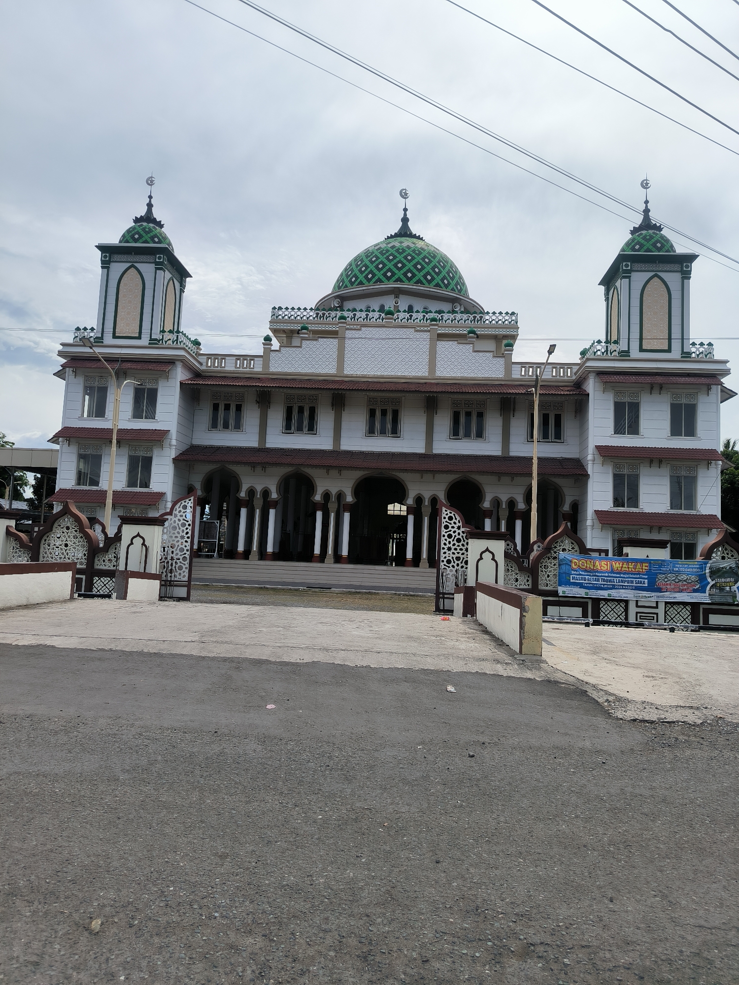 A wide-angle, eye-level outdoor shot captures the Grand Mosque Tengku Lampuk Saka in Dua Paya, Aceh, Indonesia, on an overcast day. The mosque features a prominent white facade adorned with green and white decorative patterns. A large central dome and two smaller domes atop minarets are covered in a distinctive green and white checkered design, each capped with a crescent moon and star finial. The building displays multiple arched windows and entrances, with brown-tiled roofs on its lower sections.
In the foreground, an asphalt road transitions into a concrete pathway leading to two ornate metal gates, which are integrated into a decorative fence featuring intricate white and brown Islamic-inspired patterns. A tall streetlamp stands to the left of the main entrance. Overhead, several utility wires crisscross the cloudy sky.
No people or specific activities are visible, presenting a serene architectural view. On the right side, a blue and white banner provides details for "DONASI WAKAF" (Waqf Donation) "UNTUK PEMBANGUNAN & RENOVASI RUMAH IMAM" (for the construction and renovation of the Imam's house) for "MASJID RAYA TENGKU LAMPUK SAKA." The banner also specifies its location as "JL. RAYA BANDA ACEH MEDAN KM. 13 GAMPONG DUA PAYA KEC. INGIN JAYA KAB. ACEH BESAR," confirming the geographic context. Banking details for "BANK ACEH" and contact phone numbers are also displayed on the banner.