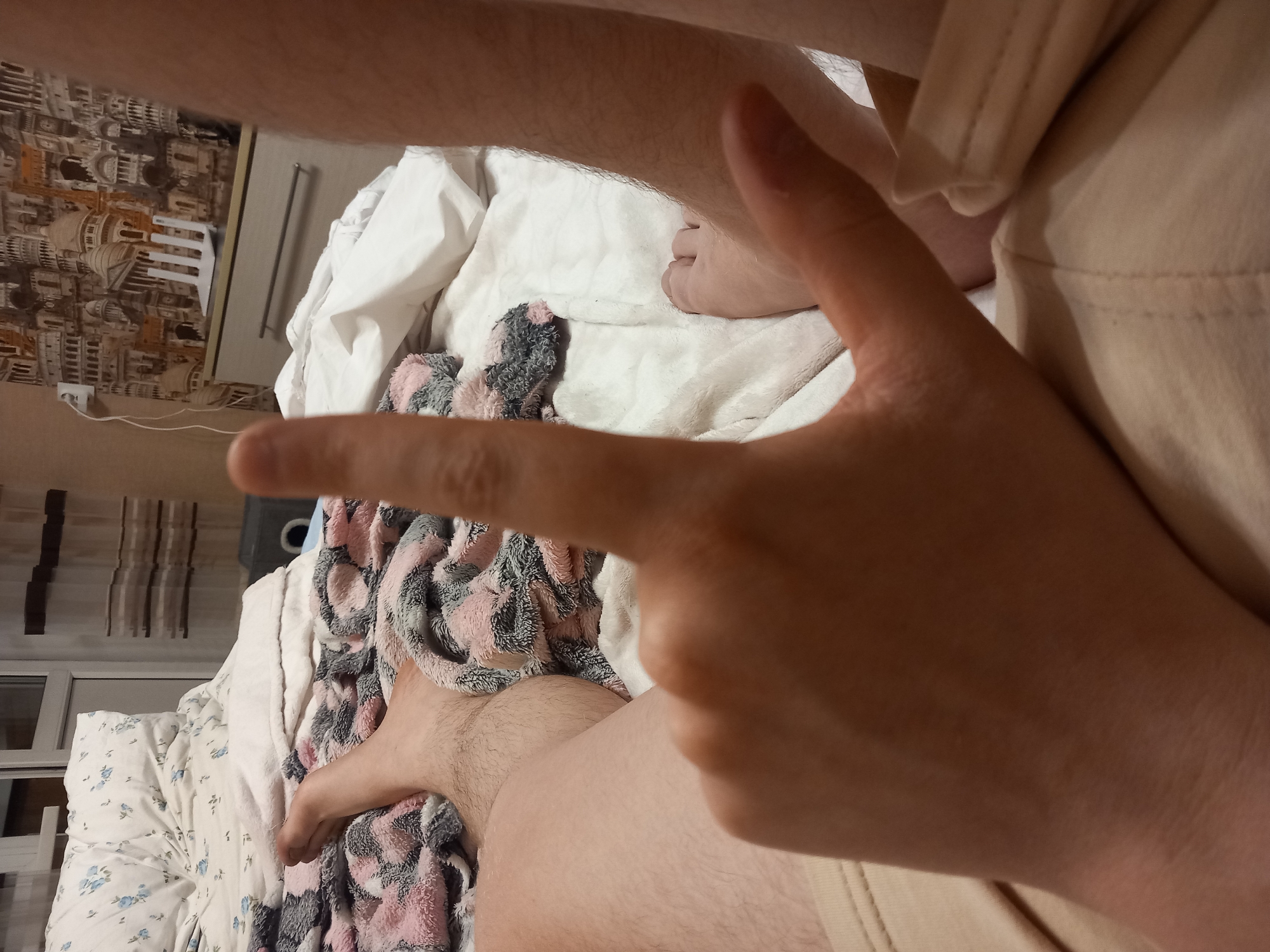 In an indoor setting, a person's hand forms a "L" shape with their index and thumb fingers extended. The hand is in the foreground, obscuring part of the image. In the background, a bed with patterned bedding and pillows is visible, along with a dresser and a wall decorated with a collage of building facades. The scene appears to be a bedroom.