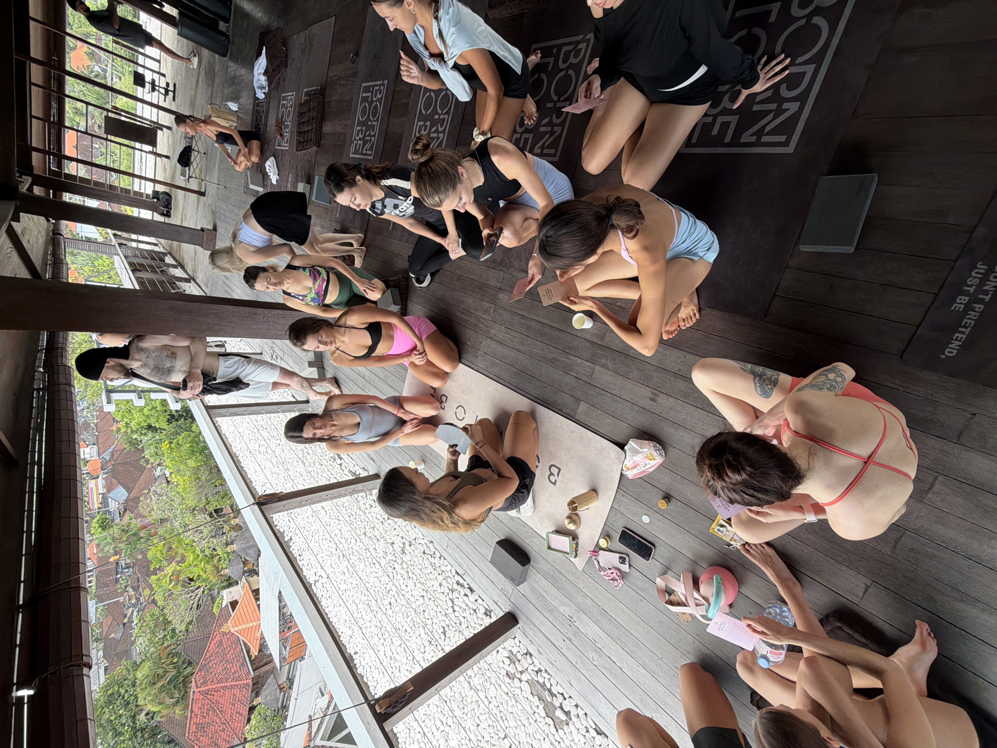 A high-angle shot shows a group of people in what appears to be a yoga or wellness retreat in Mengwi, Bali, Indonesia. The group is seated on a dark wood floor, with some individuals on yoga mats that have phrases like "BORN TO BE" and "JUST BE" printed on them. The group is engaged in an activity that involves reading from cards. Several personal items are scattered around, including phones, water bottles, small bags, and other accessories. The retreat space overlooks a lush, residential area of Bali with traditional orange-tiled roofs and dense greenery. The location has an open-air design, with wooden pillars and balustrades defining the space. The group seems to be in a post-yoga session or workshop setting.