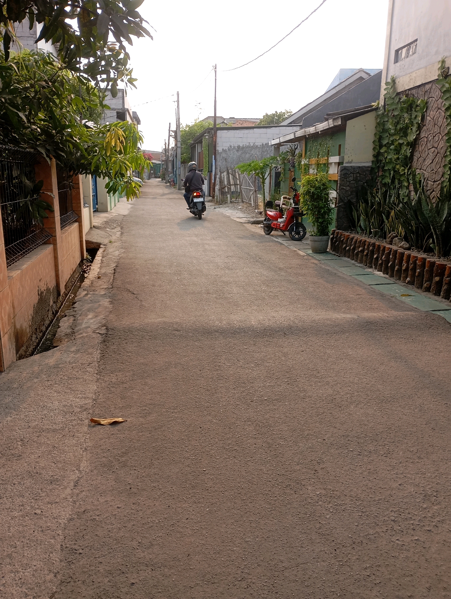 A person rides a dark-colored motorcycle down a narrow paved street in Babelan, Indonesia. The rider is wearing a gray long-sleeved shirt and a helmet. The street is flanked by residential buildings and vegetation. To the left, a terracotta-colored wall with a metal fence separates the street from a building and a small drainage ditch. To the right, a red electric scooter is parked in front of a building with green walls and a stone facade. A potted plant stands next to the scooter. The sky is overcast and the lighting suggests it is daytime.