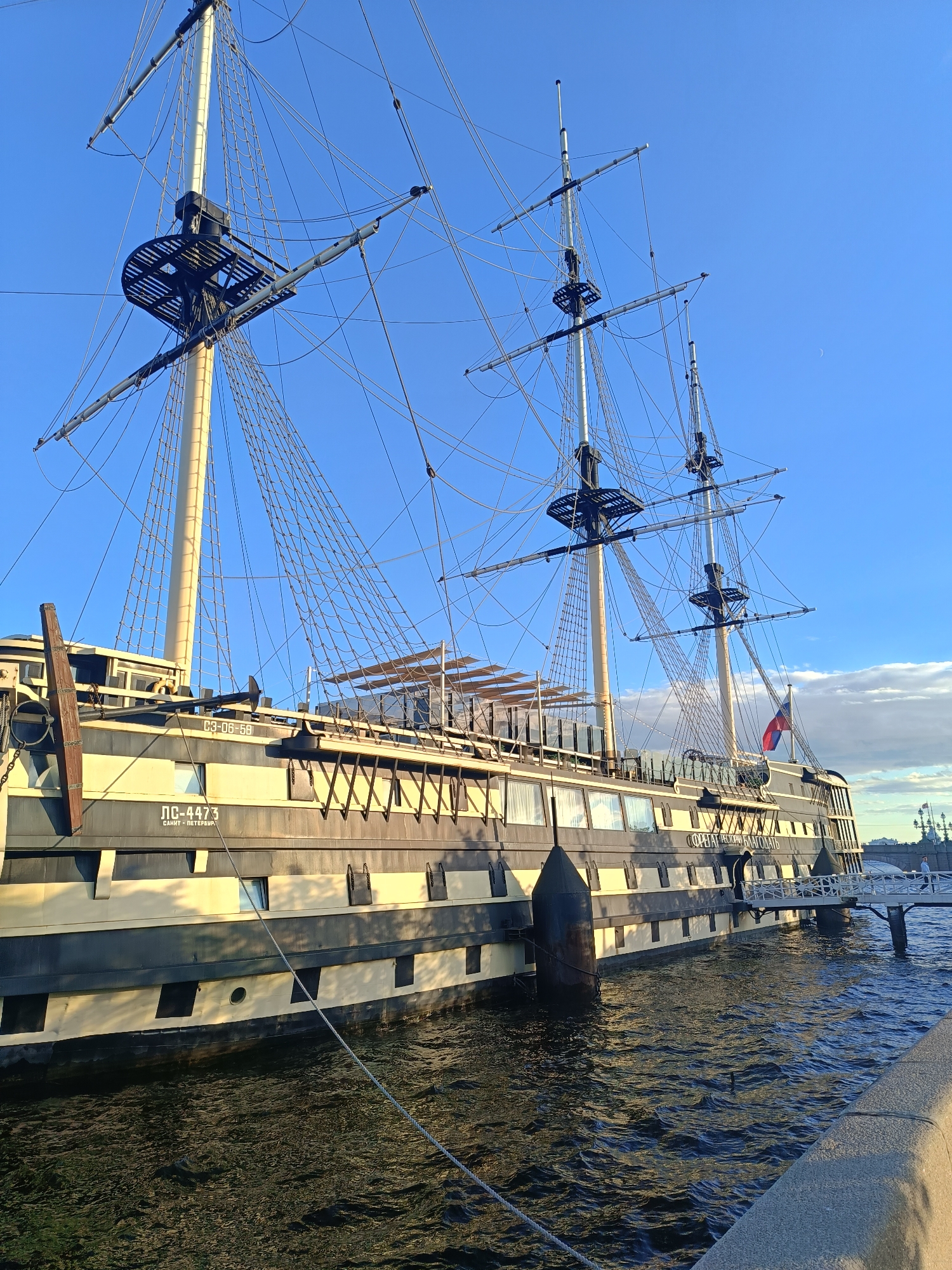 The image captures a historic sailing ship docked in the Neva River in Saint Petersburg, Russia. The ship, named "Pobedel" (Победа), is a replica of a 19th-century frigate and serves as a restaurant. Its tall masts and intricate rigging are prominent against a clear blue sky with a few wispy clouds. The ship's hull is painted in alternating dark and light stripes, and its decks are visible, with some areas covered by awnings. A gangway extends from the ship to a stone embankment, where the camera is positioned. The water of the Neva River is dark and rippled, reflecting the sunlight and the ship. In the background, parts of the Saint Petersburg skyline are visible, including a bridge and buildings. No people are clearly visible in the immediate foreground. The overall scene depicts a sunny day in a picturesque location, highlighting the grandeur of the historic ship.