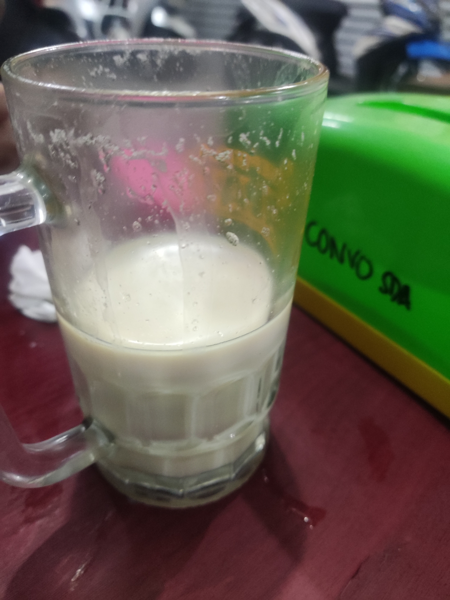 A close-up, eye-level shot shows a clear glass mug filled with a milky white liquid, likely soy milk, resting on a dark red, possibly wooden surface. The mug has a handle on the left side. The surface appears slightly wet in places, suggesting spills or condensation. To the right of the mug is a bright green object with yellow trim, possibly a box or container, with black text that reads "CONVO SDA". The background is out of focus and appears to be indoors, with hints of motorcycles and other indistinct objects, suggesting a public area or cafe. The lighting is dim. The scene is set in the city of Sidokepung, Indonesia.