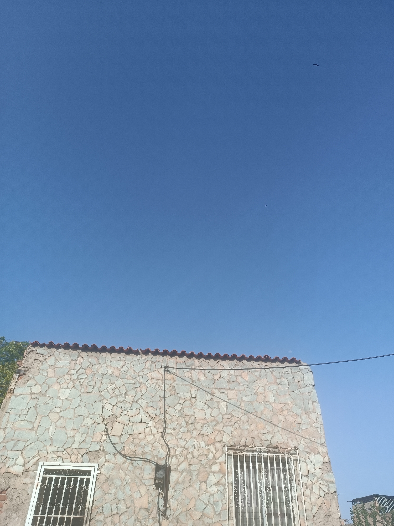A low-angle, daytime shot depicts a building exterior against a bright blue sky in Porlamar, Venezuela. The structure features a rough, light-colored stonework facade topped with a row of curved red roof tiles. Two barred windows are visible on the wall, one to the left and one to the right, both with white frames. Utility wires and a gray electrical meter are attached to the facade. Two birds are discernible in the otherwise clear blue sky. Part of a green tree is visible on the left side of the frame.