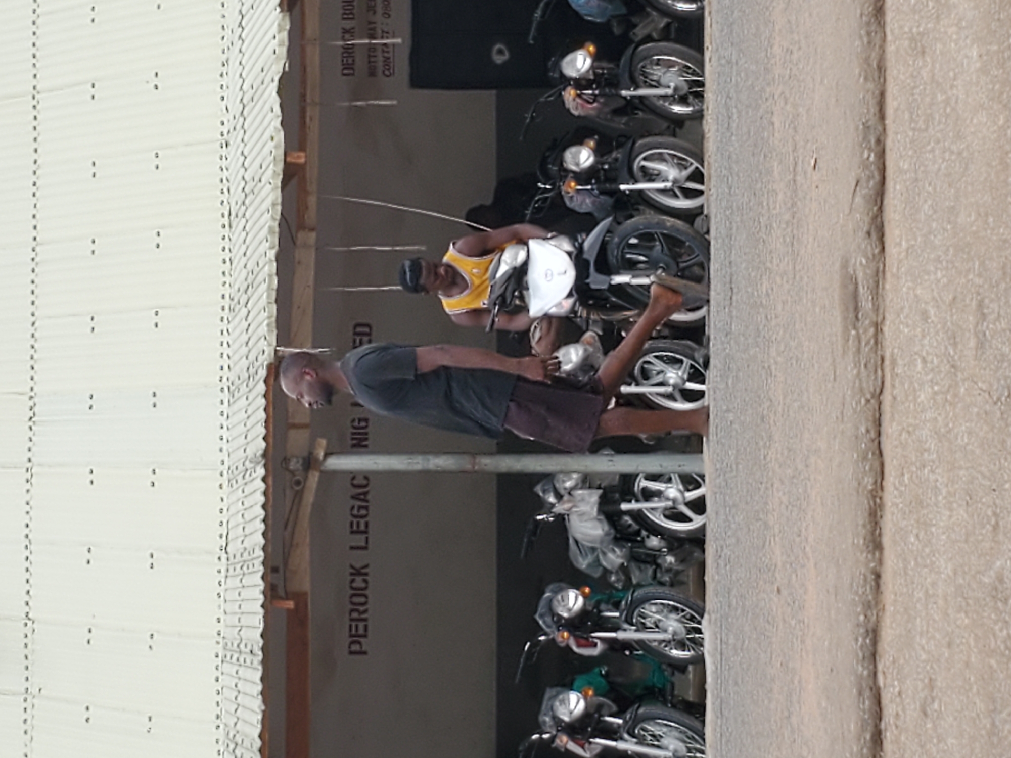 The image depicts a roadside setting in Mararaba, Nigeria, with a partial view of a building wall displaying the text "PEROCK LEGAC NIG" and "DEROCK BOL". Several motorcycles are lined up, apparently for sale. Two men are present: one is wearing a yellow sports jersey and a black cap, and the other is wearing a gray t-shirt and shorts. The wall also has the text "MOTTO MAY JE" and "CONTACT: 080". The structure on the left appears to be a corrugated metal building or shelter. A concrete wall is visible on the right side. The focus appears to be on the motorcycles and the people near them.
