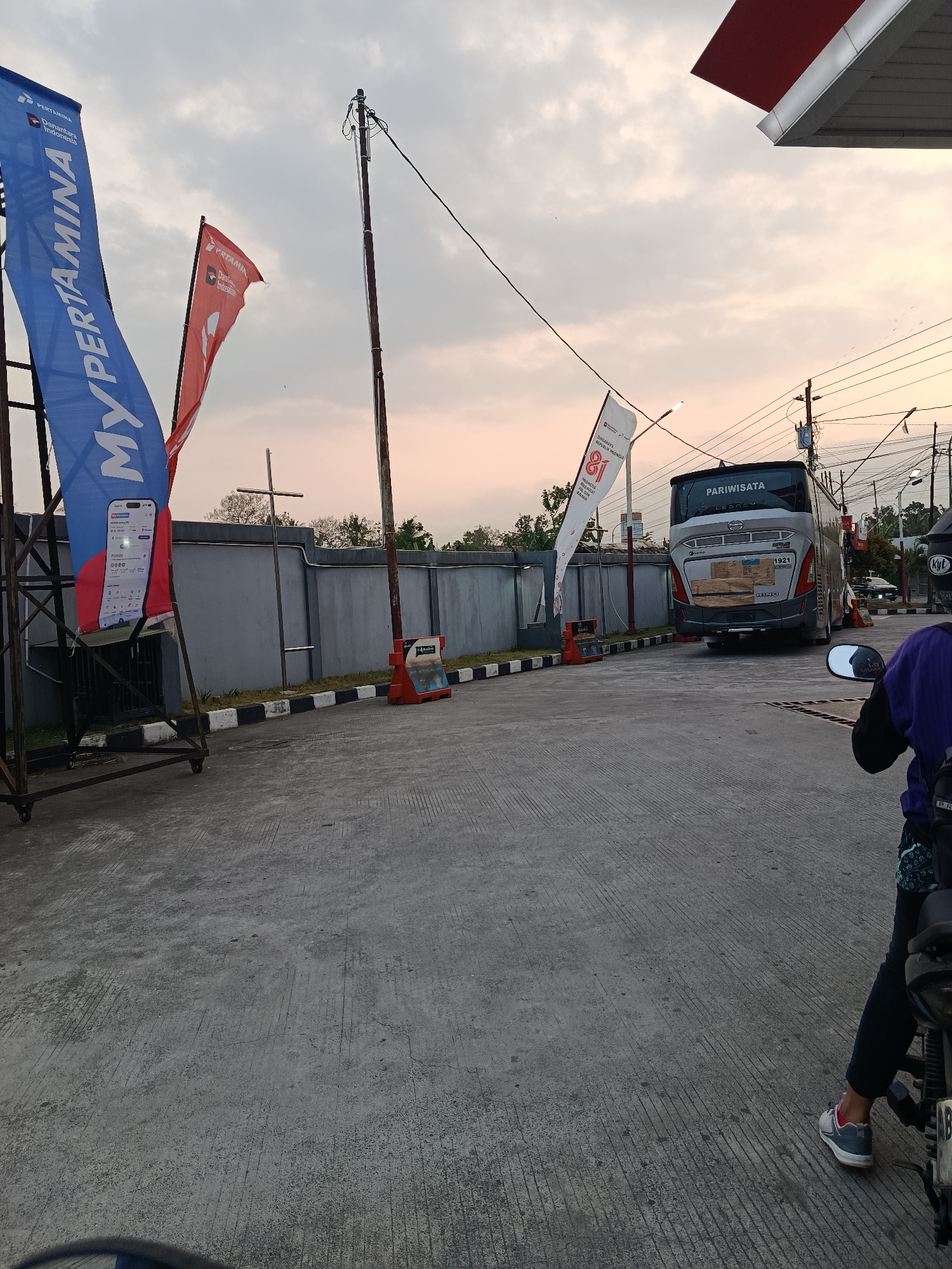 The image depicts an outdoor scene at what appears to be a Pertamina gas station in Sedayu, Indonesia. In the foreground, a person is seated on a motorcycle, viewed from the side, with their legs and feet visible. They are wearing dark pants and sneakers. To the left, a large blue banner with the text "MY PERTAMINA" and a depiction of a smartphone screen is displayed on a metal frame. A red flag with "PERTAMINA" is also visible. In the background, a silver tour bus with "PARIWISATA" written on its rear is parked. The bus has various stickers and labels on its back. The ground is paved with concrete, and a low grey wall runs along the back of the scene, with trees visible beyond it. The sky is overcast with hints of a sunset. Electrical poles and wires are present, indicating an urban or semi-urban environment.