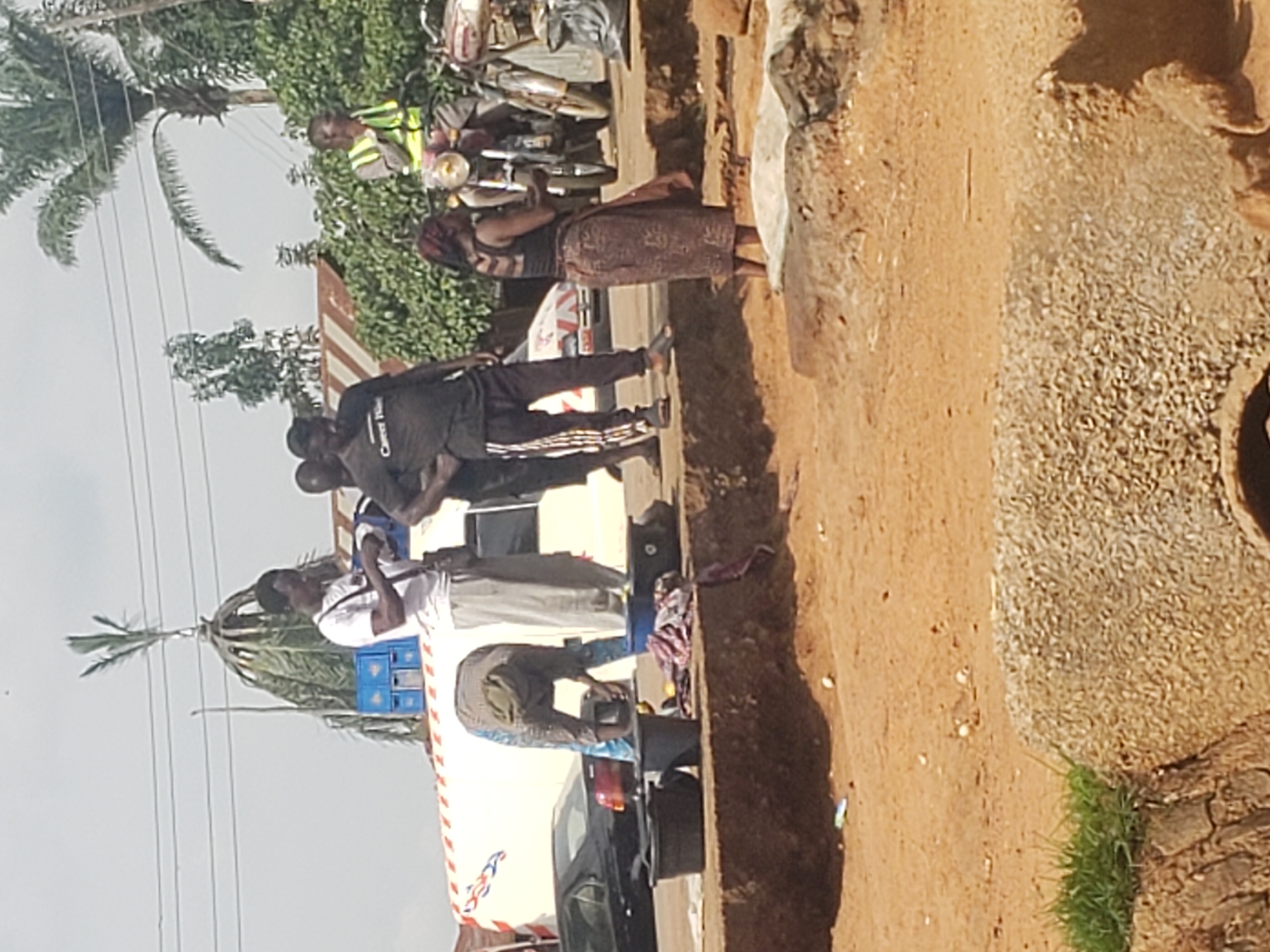 In Kachia, Nigeria, a group of individuals are gathered near a road. In the foreground, a person is bent over, appearing to wash or handle items in large black containers situated on a raised dirt embankment. Next to these containers are folded clothes. Behind this activity, a white van with red and blue markings is visible, parked on the road. Several men are standing around the van, one of whom is holding a white object. Further down the road, a man wearing a reflective yellow vest is seated on a motorcycle. Another motorcycle is parked nearby, and a woman wearing a patterned skirt and a dark top is standing next to it. The environment appears to be a street or market area, with trees and buildings visible in the background. The ground is primarily made of dirt and sand.