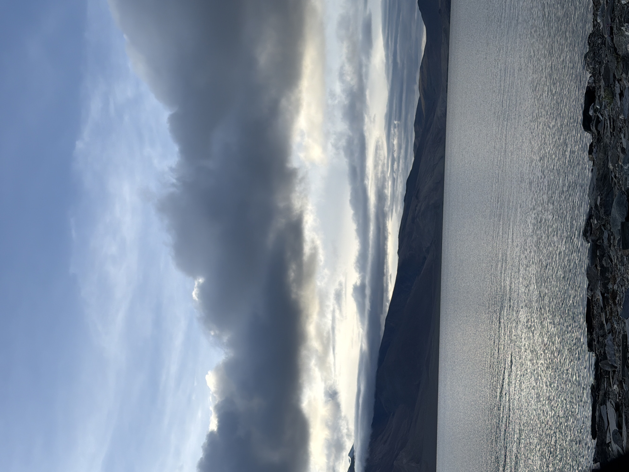 A wide shot captures a serene, rocky shore leading to a vast, tranquil lake. The lake's surface reflects the dramatic, cloudy sky above. In the background, a chain of dark, imposing mountains forms a stark silhouette against the horizon. The mountains are partially shrouded by wisps of cloud, adding to the atmospheric, somber mood of the landscape. The scene is devoid of any human presence or activity, emphasizing the raw beauty and solitude of the natural environment.