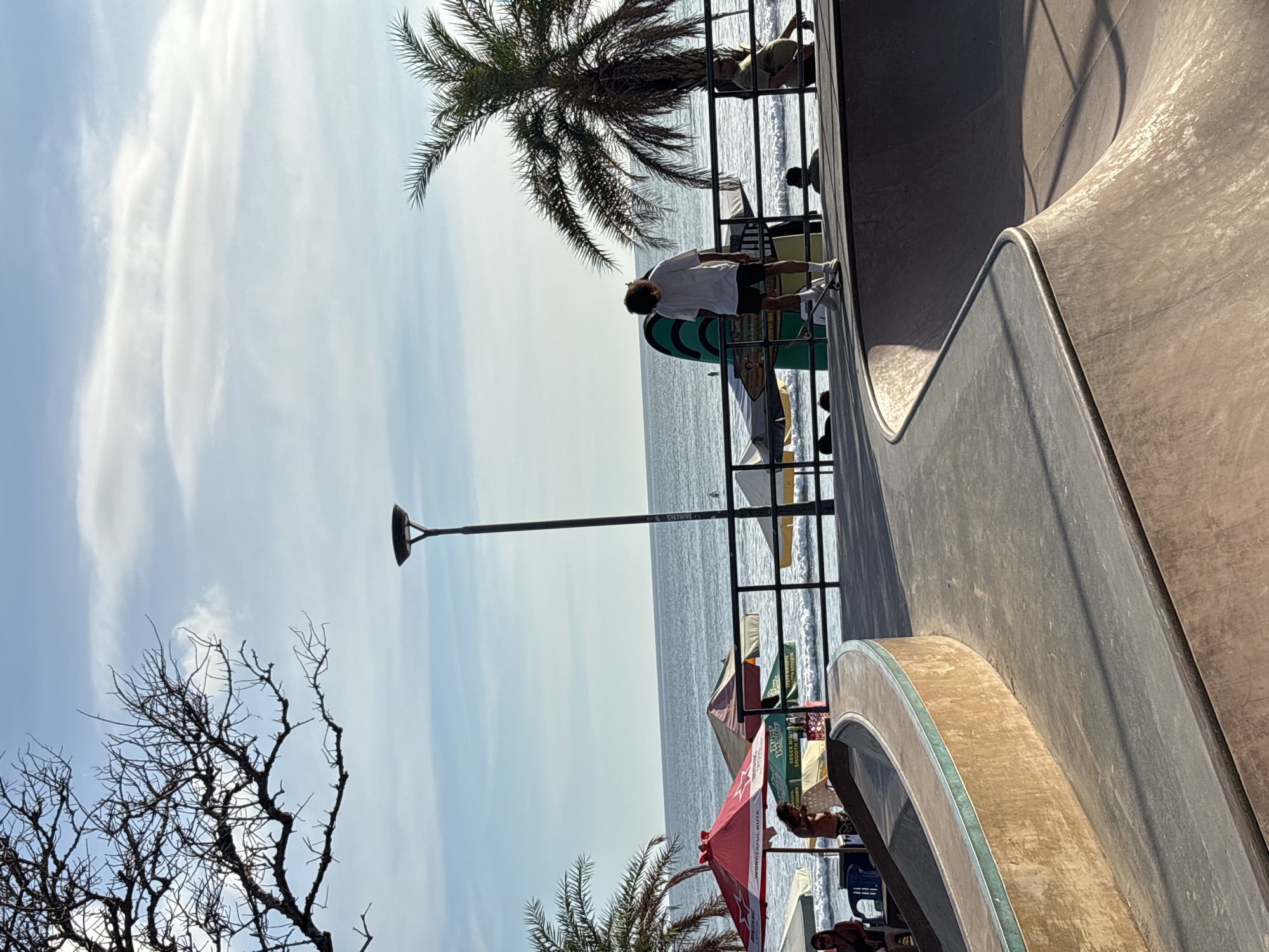 The image shows a view from a concrete skatepark towards the beach in Kuta, Indonesia. The sea stretches to the horizon under a partly cloudy sky. A palm tree is visible on the left-hand side and people can be seen standing or sitting near the beachfront, under a red and white branded umbrella. A man stands on the edge of the skatepark with a skateboard. A lamp post stands beside the skatepark.