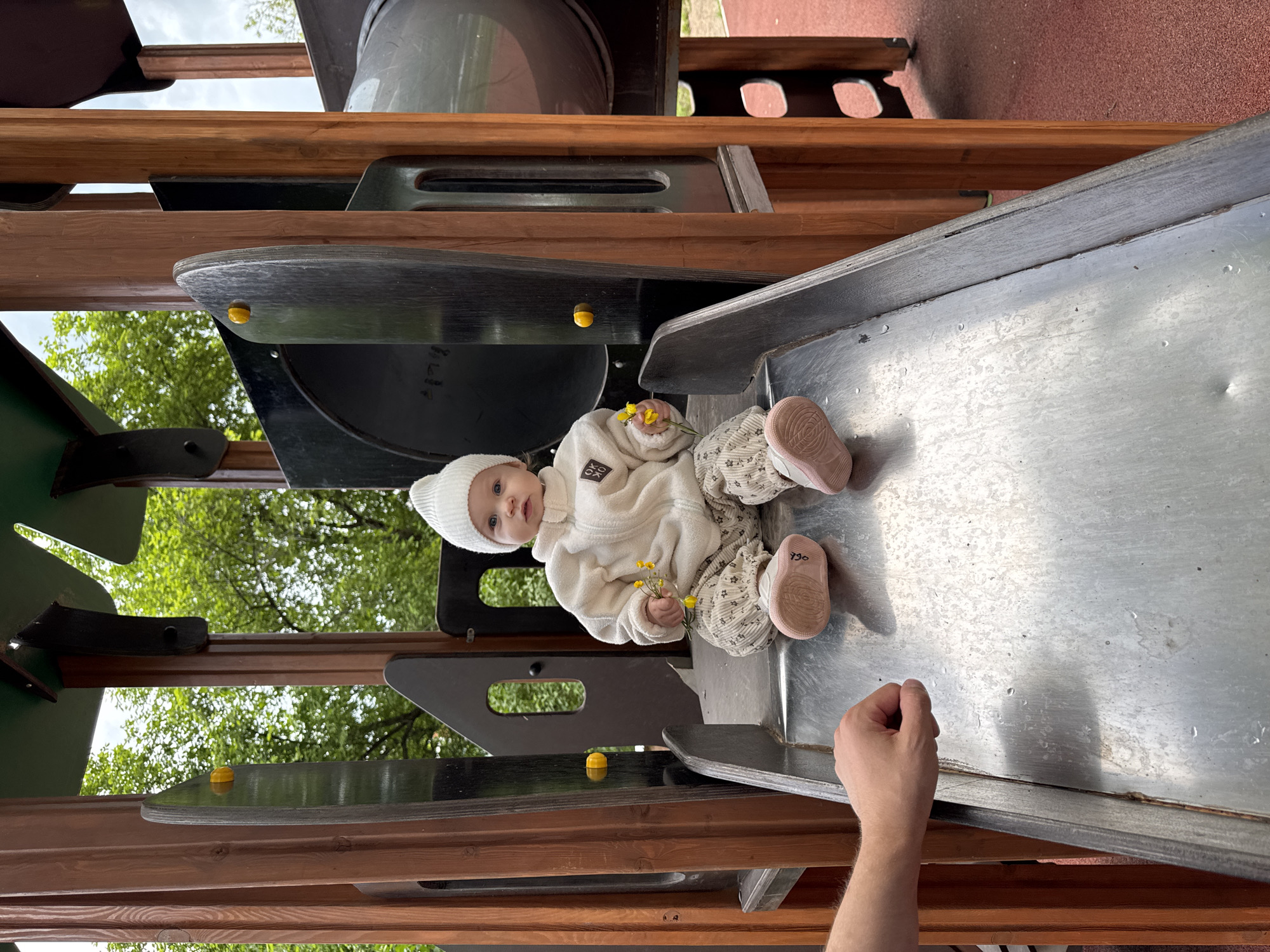 A baby wearing a white knit hat, a white sweater, and patterned pants with pink shoes sits on a metal slide. The baby is holding yellow flowers in each hand. A man's arm reaches into the frame from the bottom right corner. The slide is part of a wooden play structure with elements in green, brown, and black. Green trees are visible in the background. The location of the scene is Вассята, Russia.