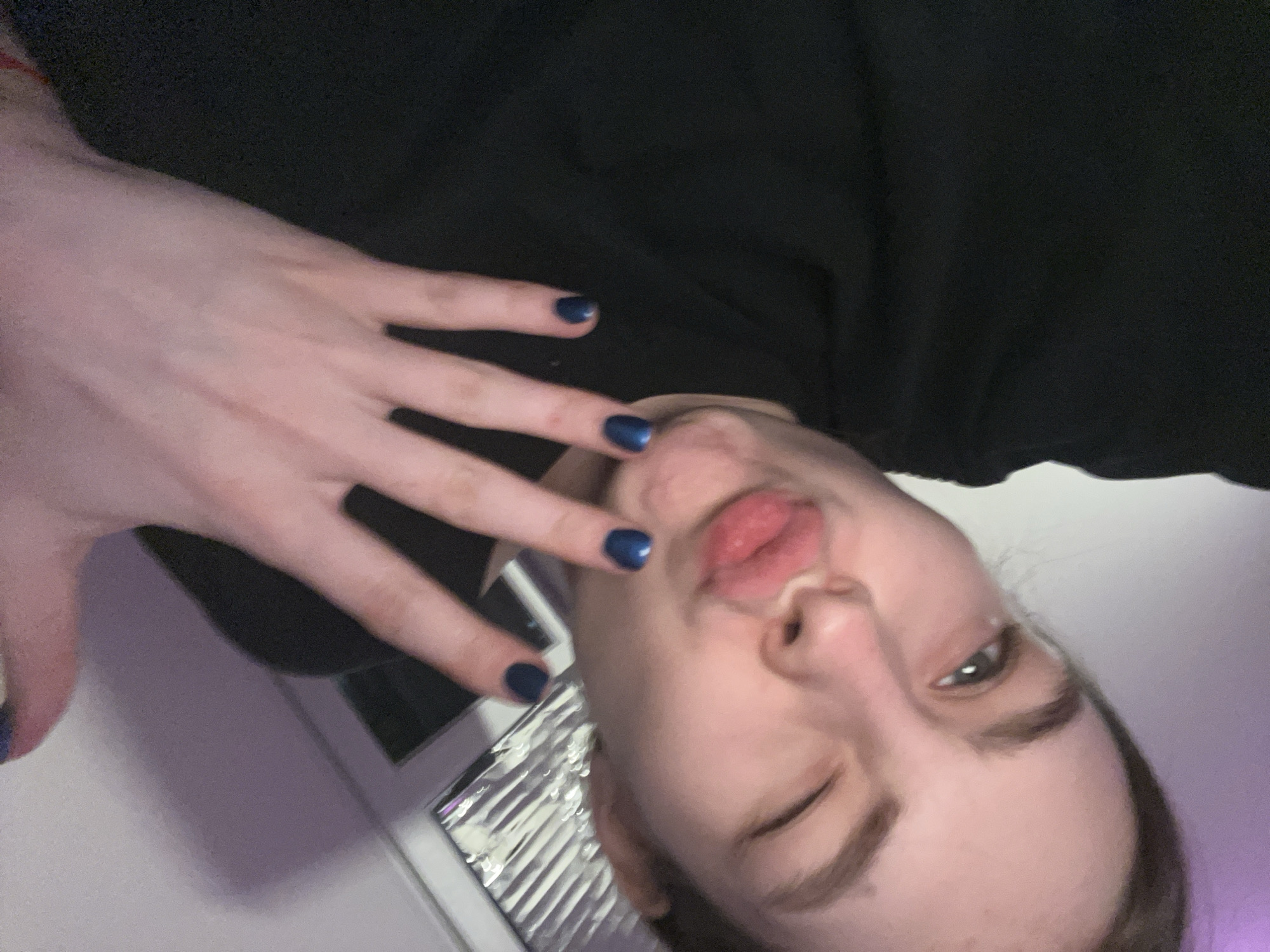 A young person poses close to the camera, with one eye closed and lips pursed. Their hand is raised, showing dark blue nail polish. They wear a dark top. In the background, there is a partially visible window with blinds and what appears to be a laptop. The location is not identifiable as Saint Petersburg, Russia, based solely on this image.