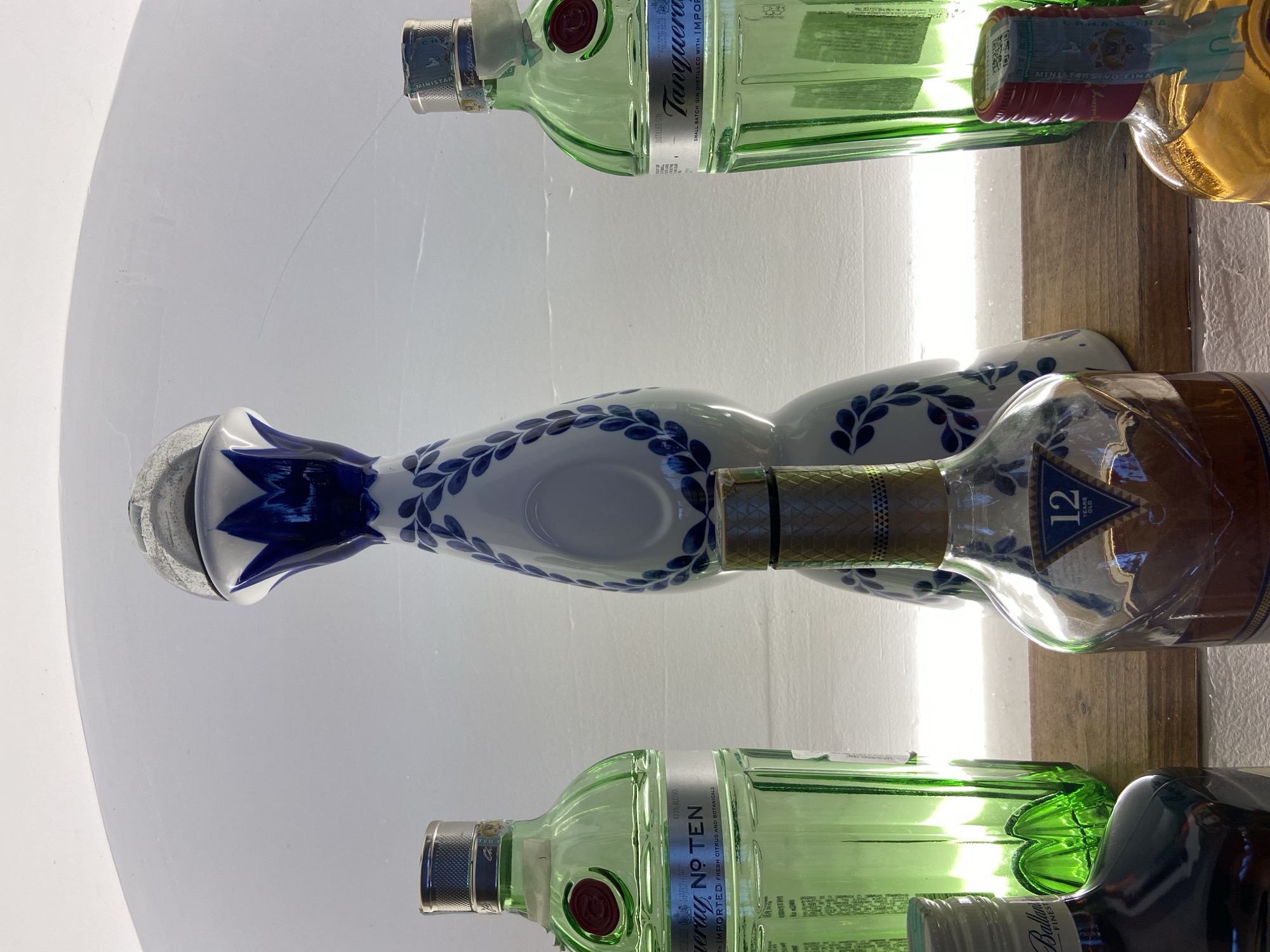 This is a close-up, eye-level shot of a shelf lined with liquor bottles. Two green glass bottles of Tanqueray gin are visible, with the labels partially obscured. Between them, there's a white, ceramic bottle decorated with blue floral patterns, next to a bottle of Ballantine's 12-year-old scotch. The background is a light-colored wall with a prominent light reflection. The photo appears to be taken in a bar or liquor store, likely in Petrovac centar, Montenegro, given that detail was requested.