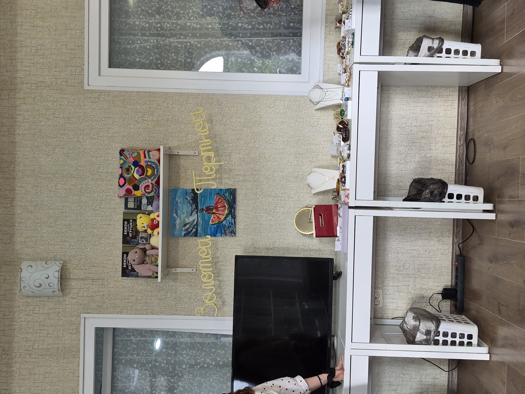 The image shows a cluttered room, possibly a living room or bedroom, with a light-colored, textured wallpaper and wooden flooring. In the background, there is a large window with a white frame and a sheer curtain featuring a delicate floral pattern.
On the left side of the image, a white wall-mounted shelf holds several items. Among them are colorful crocheted decorations, books with Russian titles such as "ВЯЖЕМ ИГРУШКИ" (We Knit Toys) and "ВЯЖЕМ" (We Knit), and two stuffed animals, a yellow teddy bear and a pink bunny. Next to the shelf, on the wall, is a decorative light fixture with a swirling pattern.
A prominent yellow, three-dimensional inscription in Russian, "Пермиева" (Permieva), hangs on the wall. To the right of this inscription is a small painting depicting a seascape with a figure in an orange dress.
A black flat-screen television is positioned in the center of the image, below the shelf and painting. Below the television, a person's arm is visible, with a black scrunchie on their wrist, suggesting they are reaching for something or resting their arm.
To the right of the television, a tall, white multi-tiered shelving unit is filled with various objects. The top shelf displays several pieces of jewelry, including rings and necklaces, presented on white busts. A small red book with "Ortodo" written on it is also visible. Further down the shelves, more jewelry is displayed, along with two white plastic baskets containing what appear to be trash bags or similar items. A power strip is mounted on the wall behind the shelves, with a black cord connected to it.
The overall impression is one of a lived-in space, possibly a creative environment, with personal belongings and decorative elements arranged in a somewhat disorganized manner. The lighting suggests it is daytime, with natural light coming from the window.