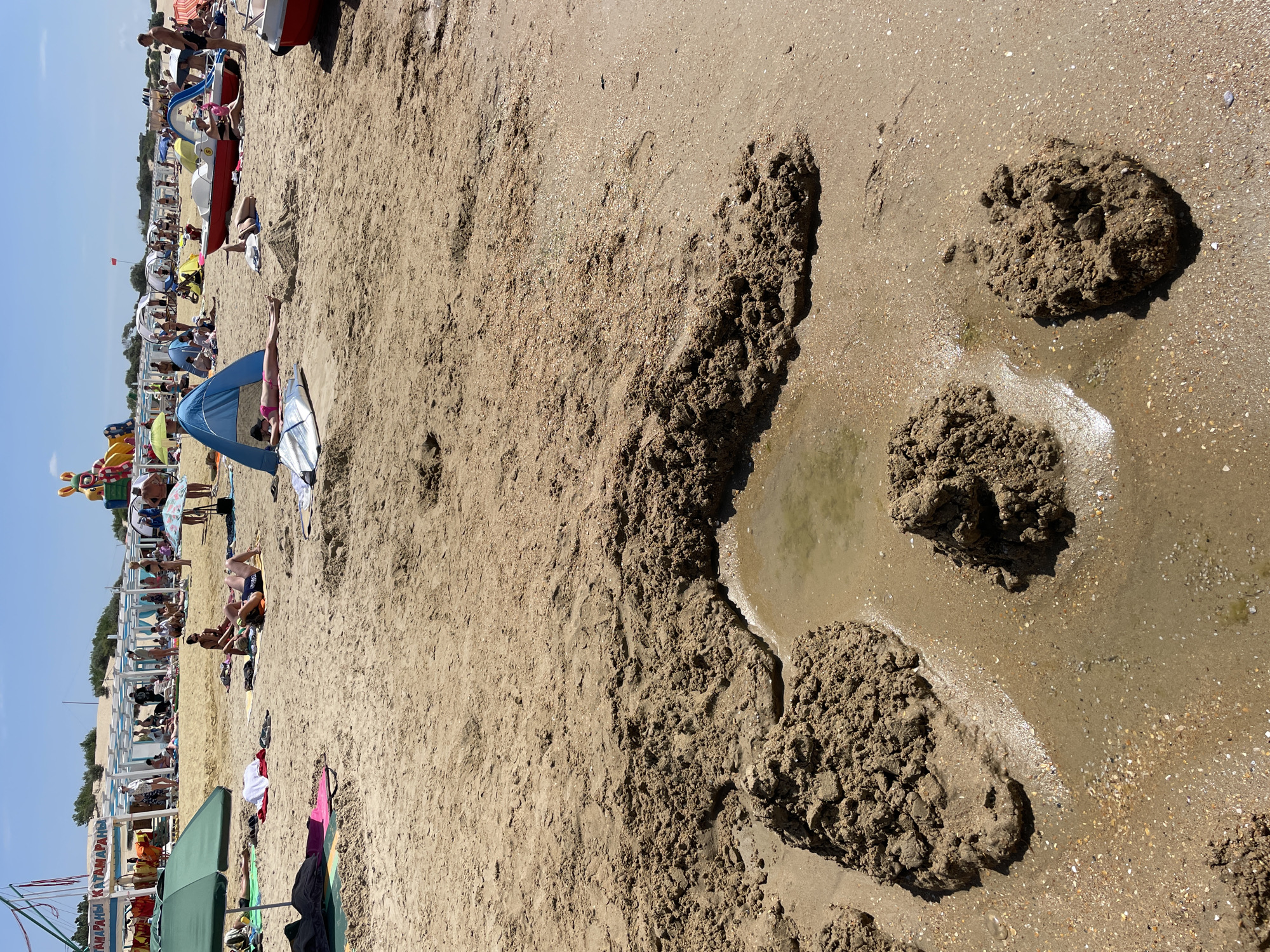 On a sunny day at the beach in Anapa, Russia, several people are sunbathing and relaxing on the sand. The beach is populated with various beach umbrellas, tents, and lounge chairs. In the background, a water park attraction with colorful slides and inflatable figures is visible, along with a building structure that appears to be a beachside cafe or service area with the Cyrillic words "КАТАМАРАНЫ" (Catamarans) and "КАТАМАРАНЫ" visible on a banner. In the foreground, several large mounds of wet sand have been piled up, with a shallow pool of water forming in the center. Some people are lying on towels, while others are sitting or walking around the beach. Two paddle boats are parked on the sand, one red and one white. The sand is speckled with small shells and debris.