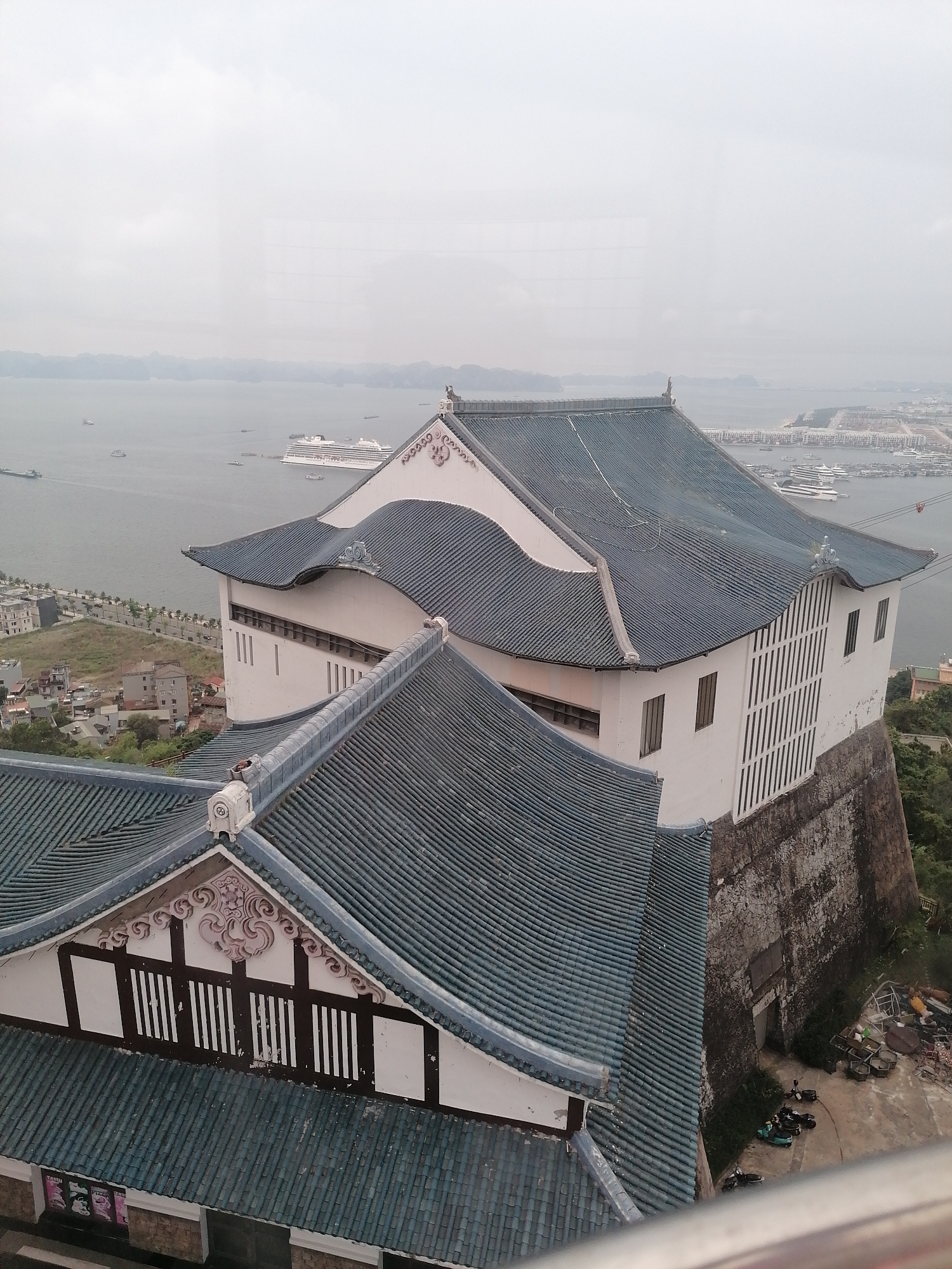 An elevated, slightly hazy, eye-level view depicts the traditional architecture of a building, likely a temple or historical structure, in Hong Gai Ward, Vietnam. The building features multi-tiered, blue-tiled roofs and white walls. In the background, Halong Bay is visible, with numerous islands and boats, including a large cruise ship. A coastal city extends along the shoreline to the right. In the foreground, below the structure, the area appears to be under construction. Scooters are parked nearby, and construction material is scattered. The sky is overcast, suggesting a humid, coastal environment.