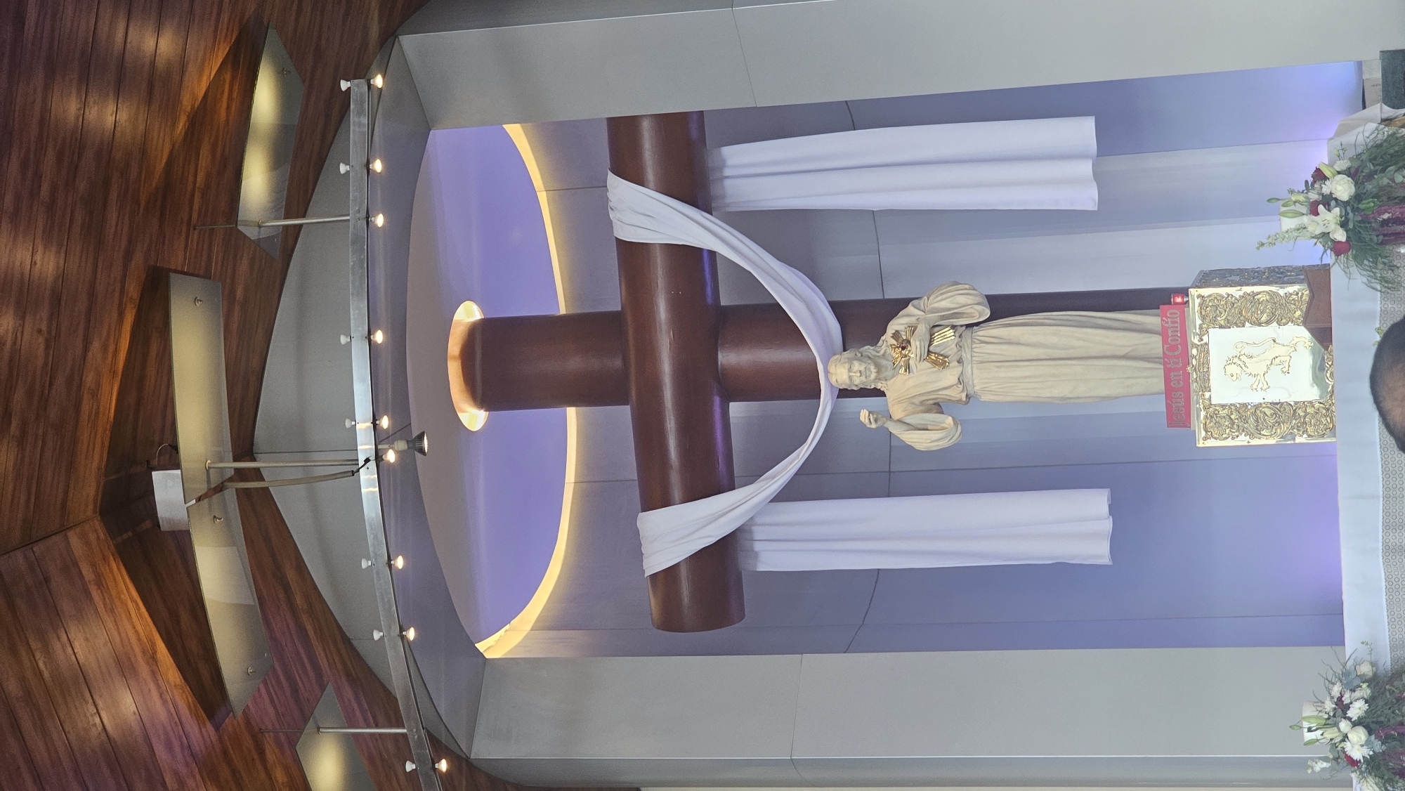 The image depicts a view of the interior of a church in San Bernardino Tlaxcalancingo, Mexico. The focal point is a large, dark wooden cross adorned with a white cloth. A statue of a saint or religious figure is affixed to the cross, appearing to be draped in additional white fabric. Adjacent to the statue is a gold-colored box or reliquary. Fresh floral arrangements flank this central scene. The surrounding architecture includes a modern design with wood paneling, steel accents, and recessed lighting that accentuates a lavender and yellow-lit curved ceiling. The room has a clean and well-maintained appearance.