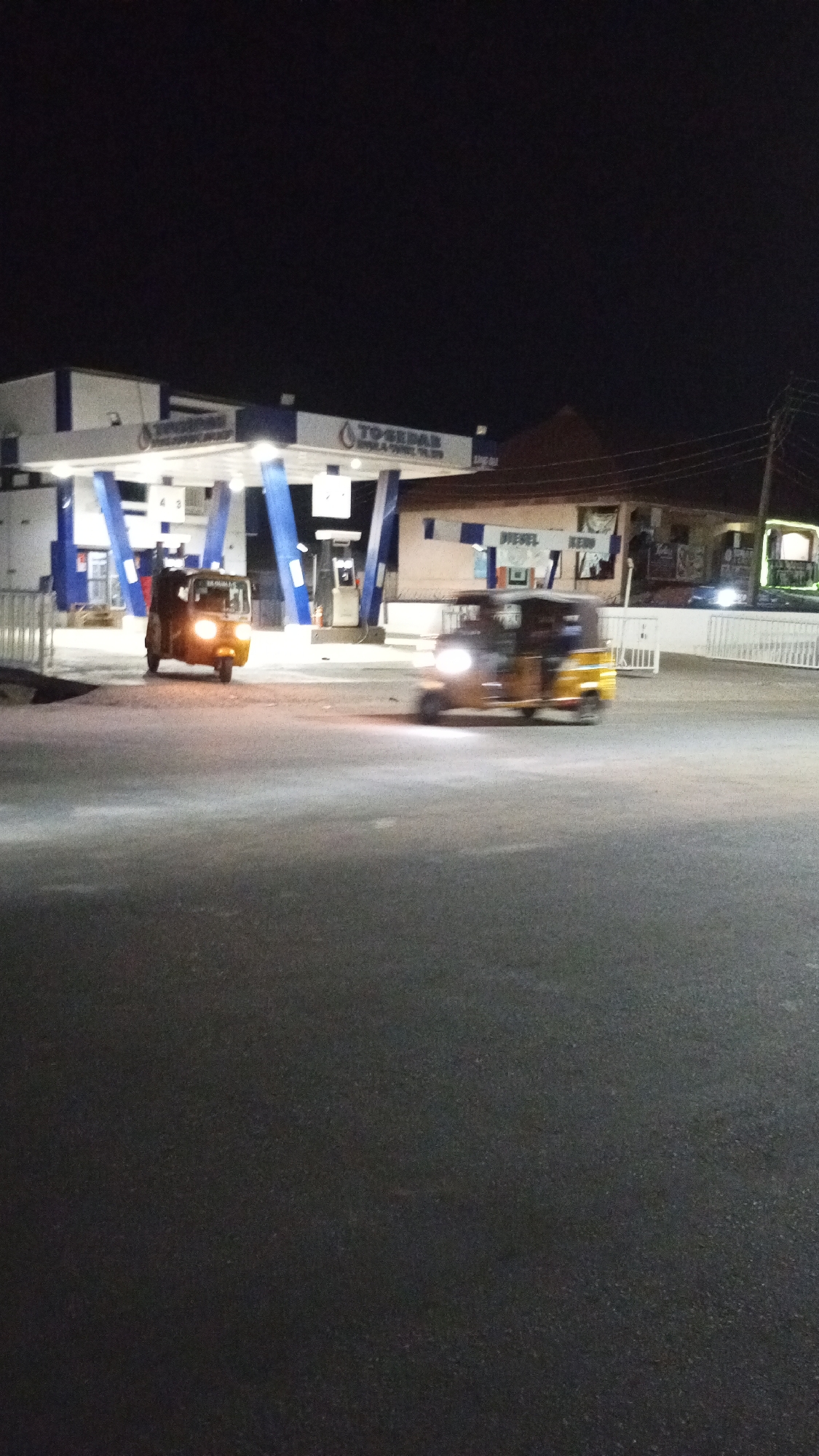 At night, the image shows a TosseDar petrol station in Adankolo, Nigeria. Two motorized tricycles or "Keke Napep" are visible. One is stationary at the petrol station. The other tricycle is blurred, suggesting motion. The surrounding area includes other buildings and utility poles.