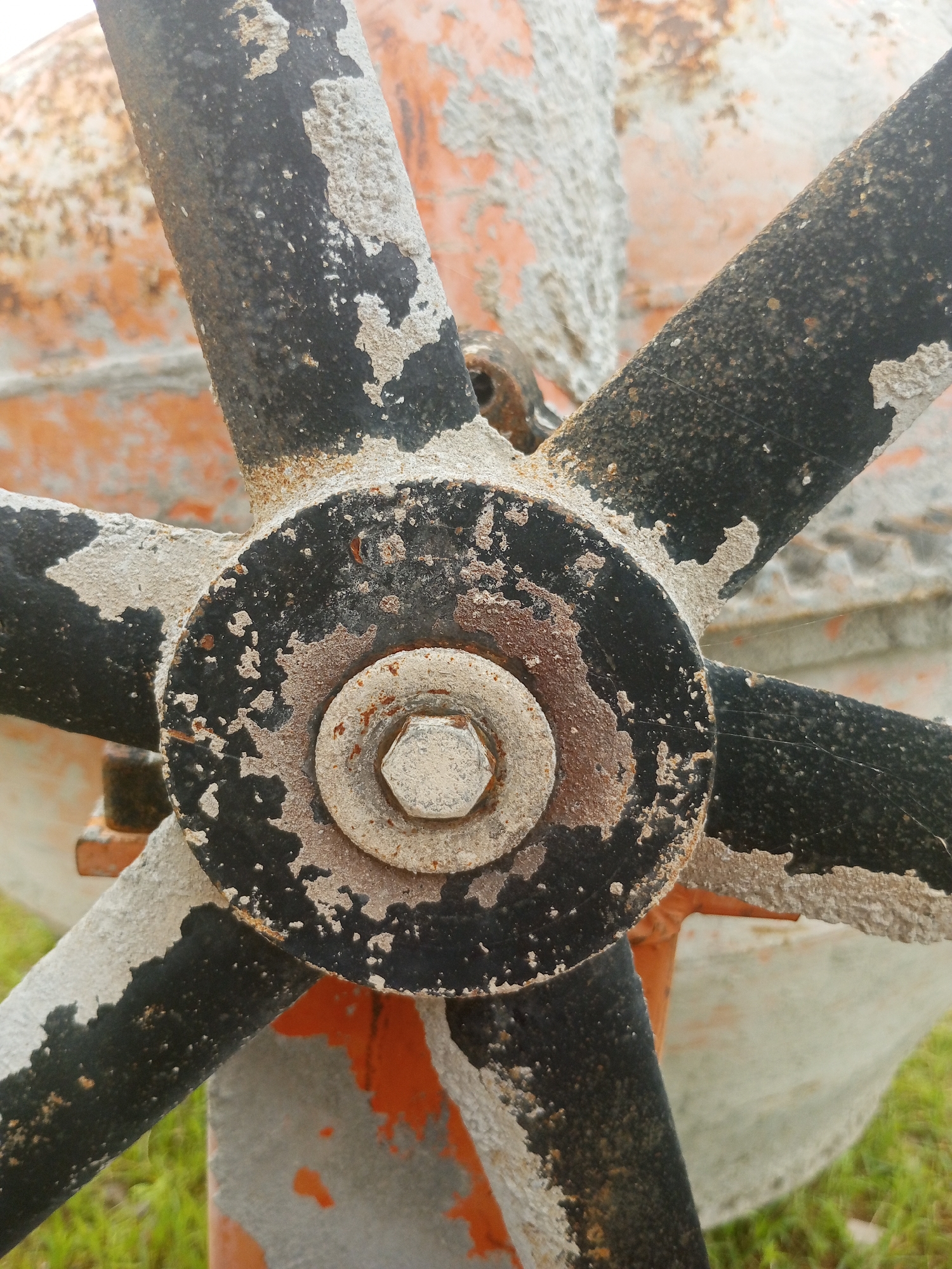 The image is a close-up, high-angle shot of a weathered, orange and black industrial object, likely a cement mixer. The central subject is a bolted metal hub with four black spokes extending outwards. The hub and spokes are heavily encrusted with dried concrete or cement, giving them a rough, textured appearance and obscuring some of the original paint. Rust is visible on the metal, particularly around the bolt and on the edges of the spokes. The background is blurred but shows hints of more orange and light-colored surfaces, also speckled with concrete. The ground beneath the object appears to be green grass. The lighting suggests it is daytime, and the overall impression is one of heavy use and exposure to the elements. There are no people or discernible text visible in the image. The setting is outdoors, with the presence of grass indicating a natural environment, but the industrial nature of the object suggests a construction site or a similar operational area. The lack of distinct features in the background makes it difficult to pinpoint a more specific location beyond a general outdoor setting.