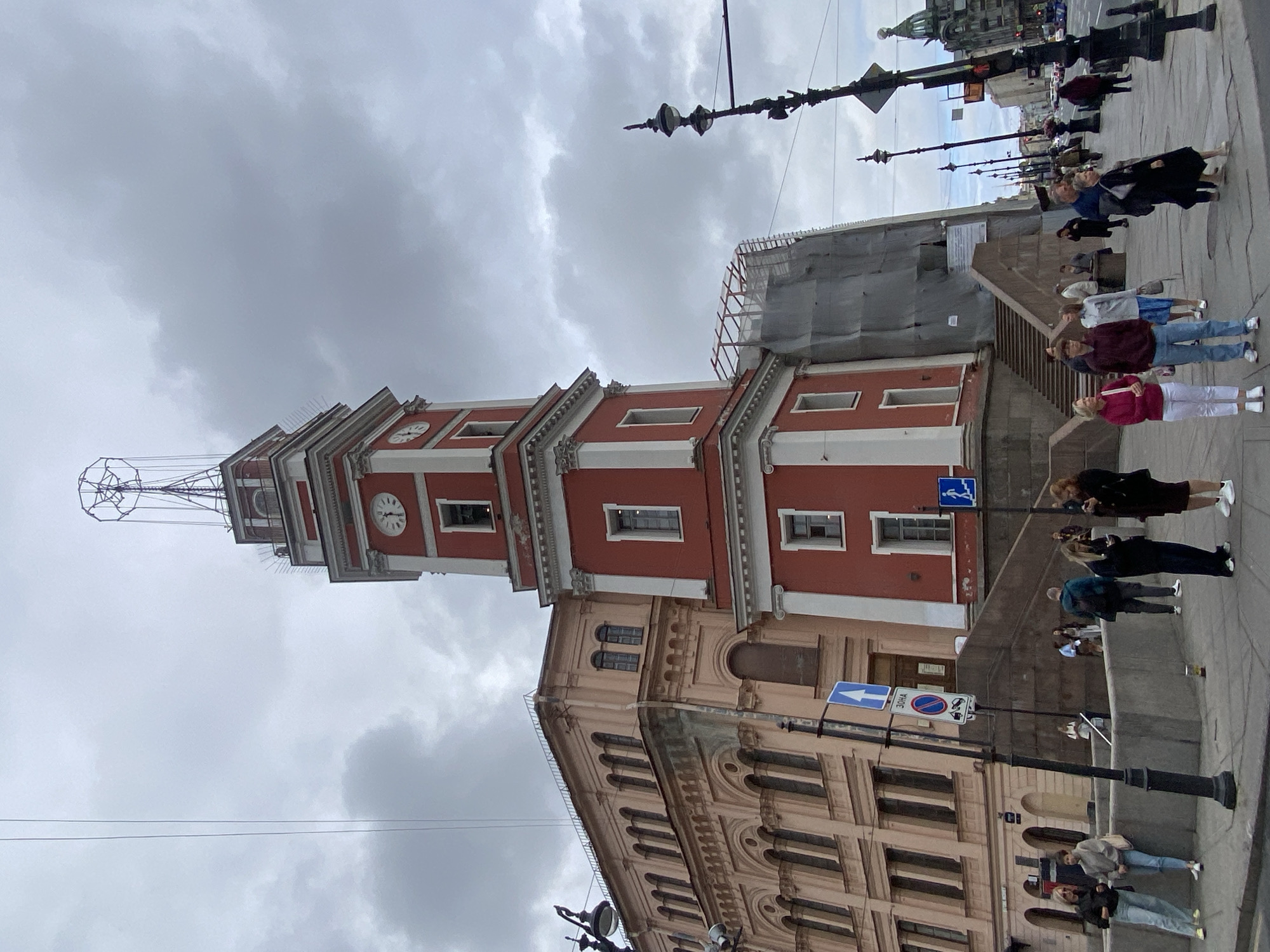 The image shows a street scene in Saint Petersburg, Russia, featuring the Singer House (Dom Knigi) and a prominent red and white tower with a clock. The sky is overcast with gray clouds.
In the foreground, a group of people are standing on a sidewalk. To the left, two women are casually dressed, one in jeans and a black jacket, the other in a gray hoodie and light-colored pants. Further to the right, a young man in a maroon hoodie and jeans stands with a woman in a pink jacket and white pants. Several other individuals are scattered throughout the scene, some looking at their phones. A black lamppost with multiple lights stands in the foreground, with traffic signs mounted on it.
The Singer House, a distinct architectural landmark, is visible on the left. It is an ornate building with arched windows and light pink facade. Adjacent to it, and dominating the center of the image, is the red tower of the Singer House, adorned with white trim and clocks. The tower is topped with a metallic structure. Part of the tower and an adjacent building are covered in scaffolding and gray tarps, suggesting ongoing construction or renovation.
A wide staircase leads up towards the red tower. On the sidewalk, a few more people are walking or standing. Beyond the immediate area of the Singer House, a street with traffic and other buildings is visible in the distance, including a green-domed building characteristic of Saint Petersburg's architecture. The overall impression is a busy urban environment on a cloudy day.
