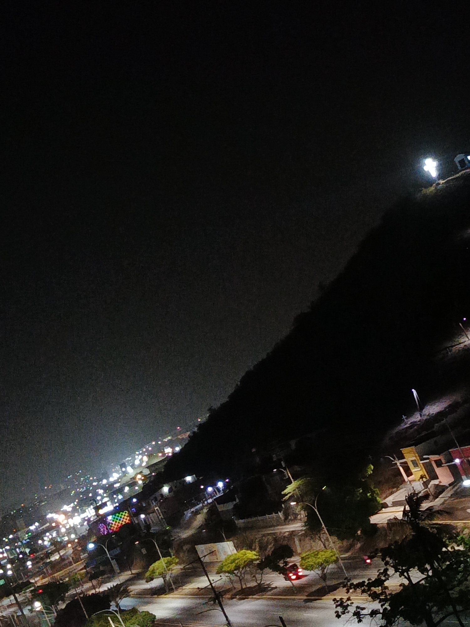 The image shows a night view of Los Robles, Venezuela. In the lower portion of the frame, city lights and illuminated streets are visible, with scattered trees and a few cars. A dark, elevated landmass, possibly a hill or small mountain, extends diagonally across the mid-ground, partially obscuring the city. At the peak of this landform, a brightly lit cross is visible. The upper part of the image is dominated by a dark night sky.