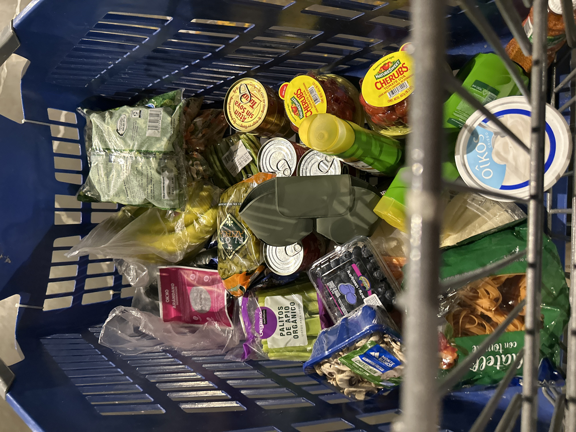 The image shows a blue shopping basket filled with groceries. Items visible include: Oikos Greek yogurt, Cherubs grape tomatoes, canned tuna, a container of blueberries, packaged mushrooms ("Monte Blanco Champiñon"), organic celery sticks ("Palitos de Apio Organico"), dried cranberries ("Okko Arandano"), a bag of bananas, green beans, packaged spinach, and a bag of pasta ("Chatel con tomate"). The OCR data includes text in Spanish. The items suggest the location could be a supermarket in an area with a Spanish-speaking population. While the OCR data contains "Huixquilucan de Degollado, Mexico" it is not explicitly stated as the location of the grocery store in the image itself, so that has not been included.