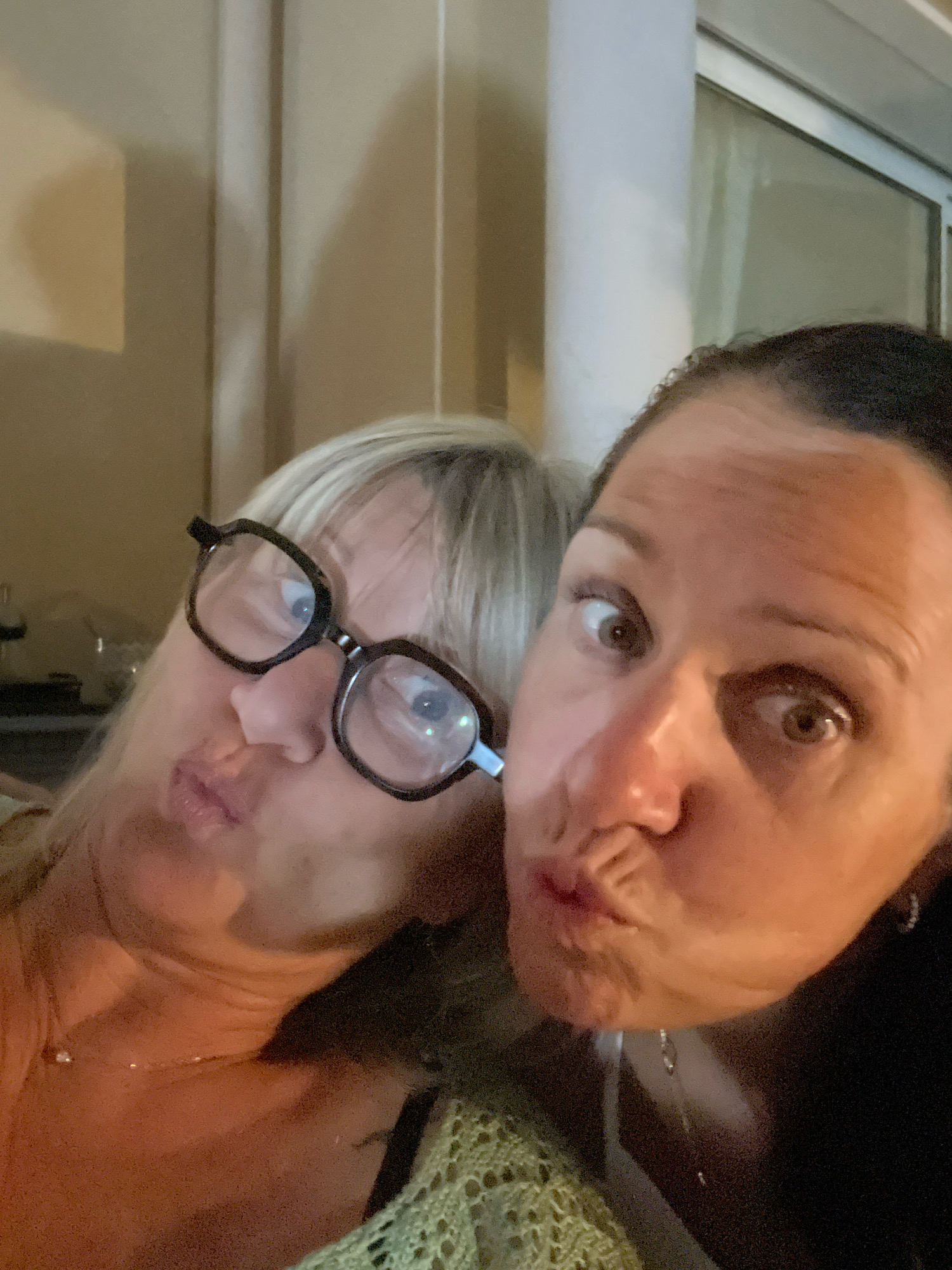 This is a selfie of two women taken indoors, possibly at night. The woman on the left has blonde hair, glasses, and is puckering her lips. The woman on the right has dark hair and is also puckering her lips, with wide eyes. They are both looking at the camera. The background appears to be an indoor setting with neutral-colored walls and a pillar. There is no indication of the specific location within Saint-Galmier, France, in the image itself.