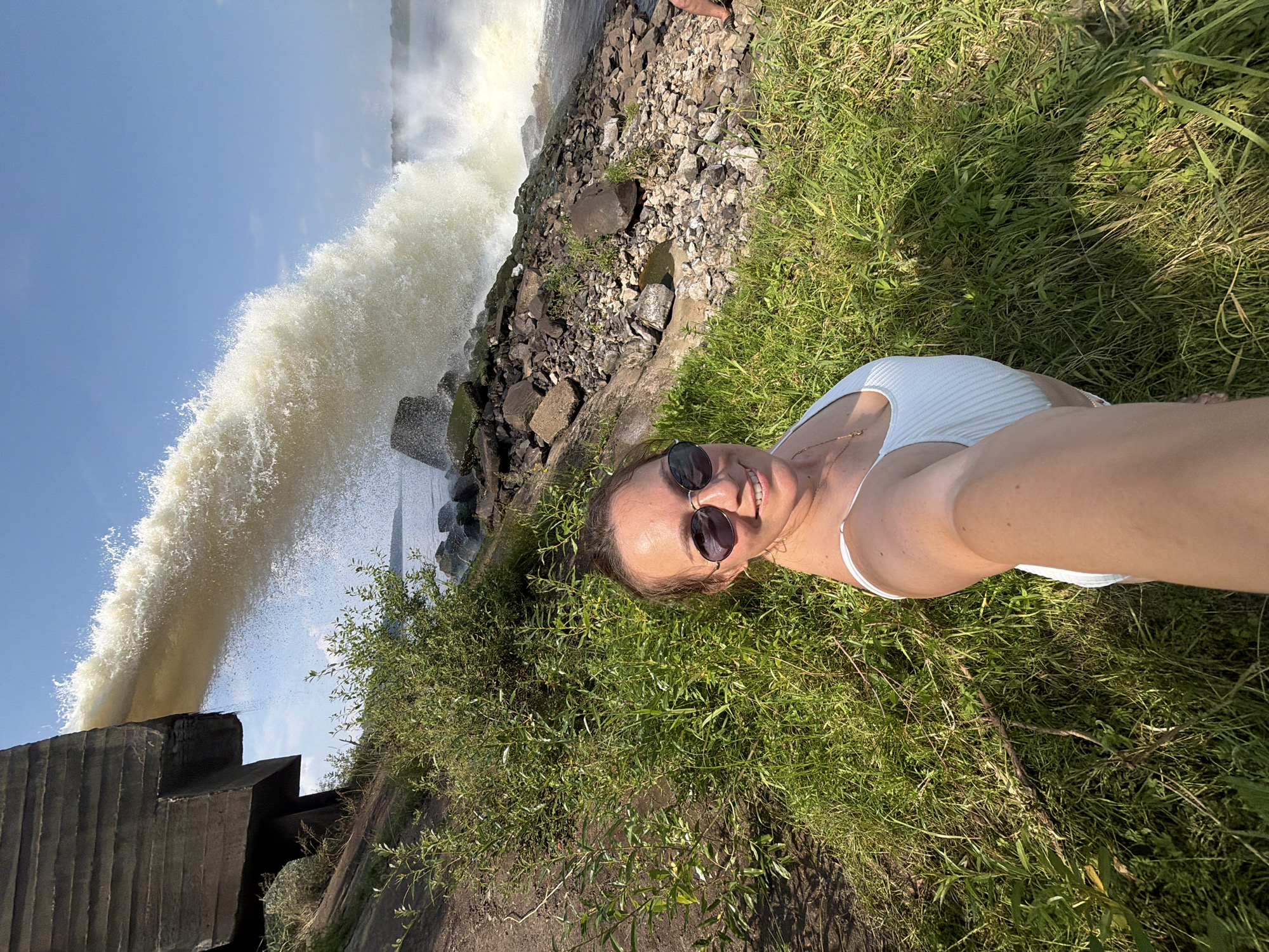 In Chernushka, Russia, a woman with sunglasses and a white swimsuit takes a selfie in front of a large concrete structure releasing a powerful stream of water into a rocky riverbed. Lush green grass surrounds the woman, who is smiling and looking at the camera. The water spray creates a dramatic effect against the clear blue sky.