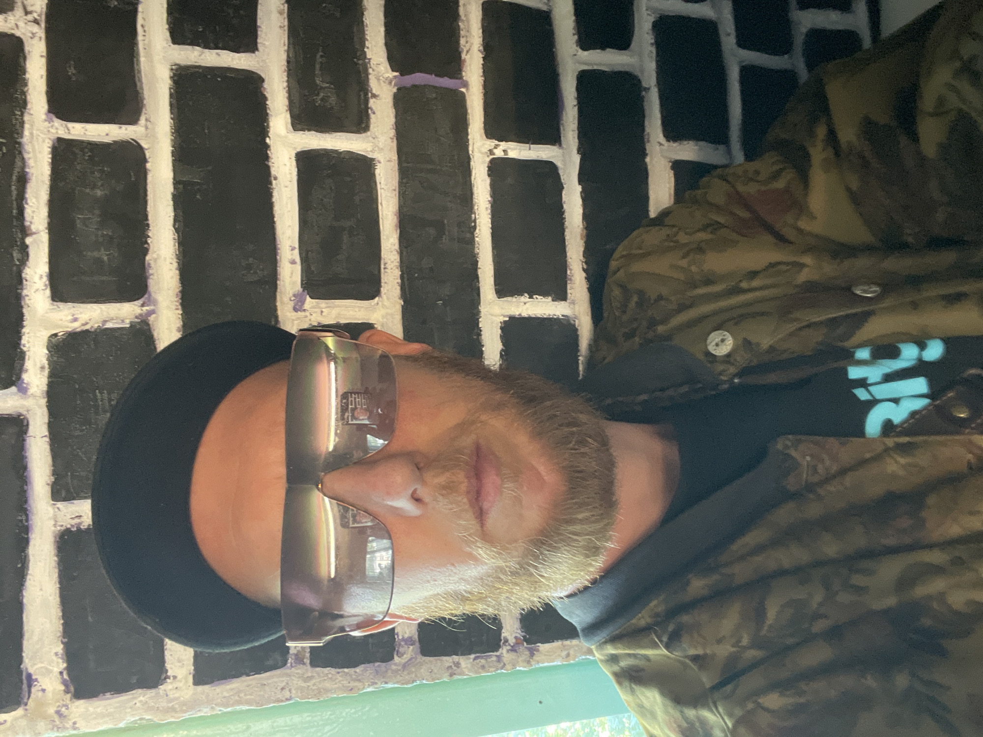 This media file is a portrait of a person with a beard, wearing sunglasses and a camouflage jacket over a black t-shirt. The person is looking down, with their eyes closed. The background is a brick wall painted black with white mortar. There is a hint of a green surface at the bottom of the frame. The lighting suggests it is daytime, likely indoors. The overall mood appears relaxed or contemplative. There is no visible text in the image, except for what might be a logo on the t-shirt, which is partially obscured. The context is Saint Petersburg, Russia.