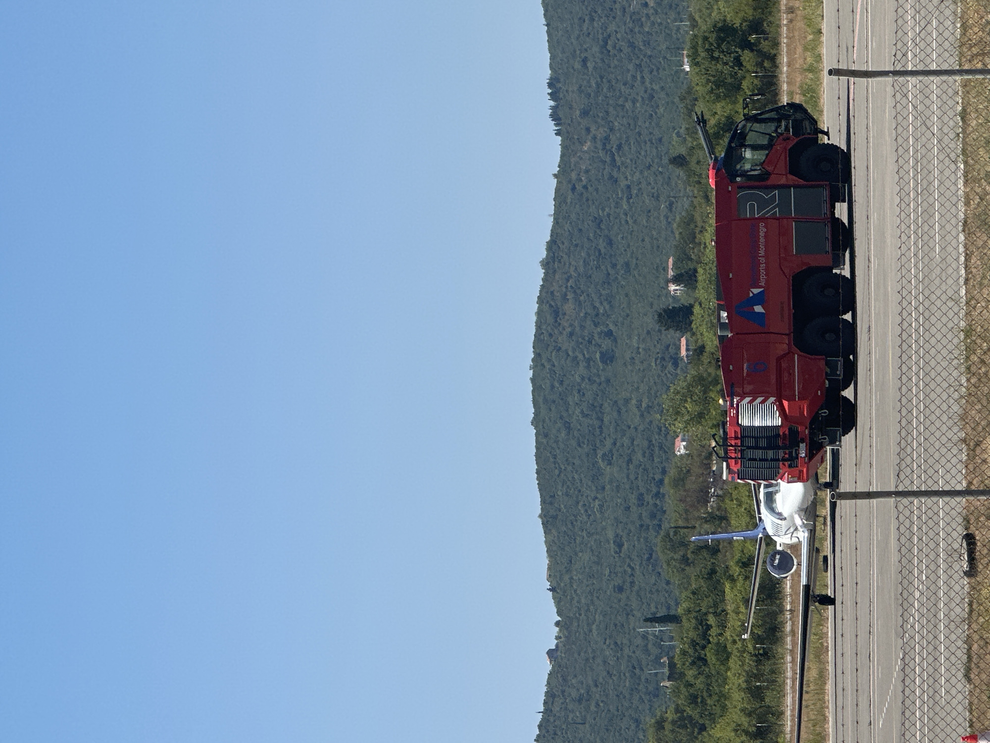 In Mrčevac, Montenegro, a red fire truck with the logo "Airports of Montenegro" is parked on an airport tarmac. To the left of the fire truck is a white and blue private jet. In the background, a lush green hillside with scattered houses is visible under a clear blue sky. The scene appears to be on a sunny day.