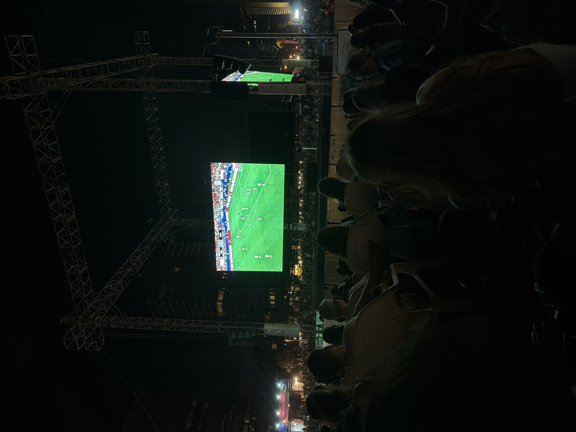 In Tel-Aviv, Israel, an outdoor screening of a football match is underway at night. A large LED screen displays the game, with players in red and white uniforms visible on the bright green field. Spectators are seated in rows of plastic chairs, facing the screen, with their backs to the camera. Some individuals are looking at the screen, while others are engaged in conversation. The scene is illuminated by the screen's glow and some ambient city lights, with tall buildings in the background. Large metal structures, likely part of the stage or rigging for the screen, are visible above the audience. A smaller screen is mounted to the right, also showing the match. The event appears to be well-attended, with a dense crowd visible beyond the seated audience.