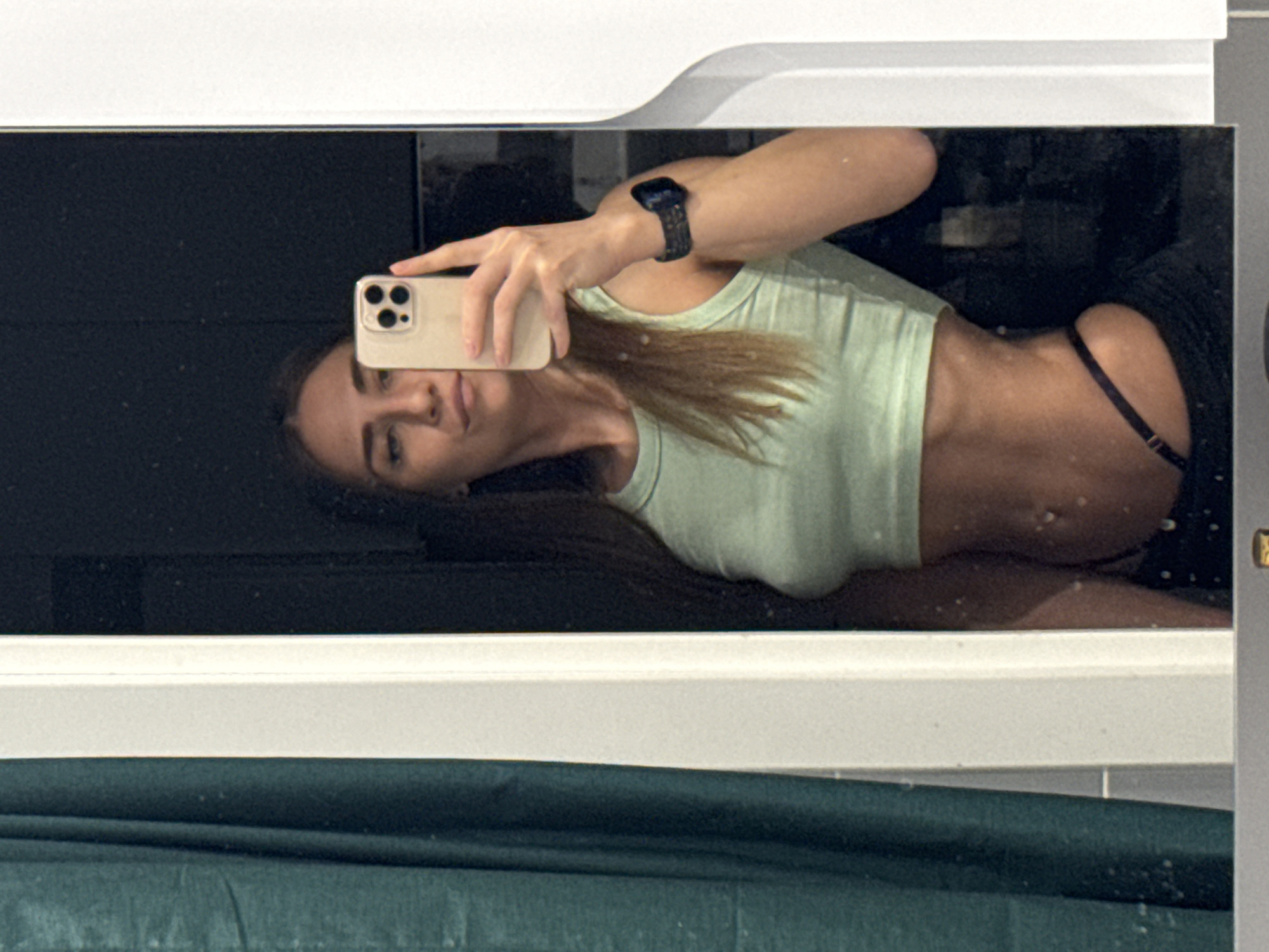 A woman is taking a selfie in a mirror. She is holding a smartphone in her left hand and wearing a light green crop top. A black watch is on her left wrist. Part of a black garment is visible on her left side and a dark green fabric is in the foreground. The location is likely indoors. There is no information to confirm the location is Lyubertsy, Russia.