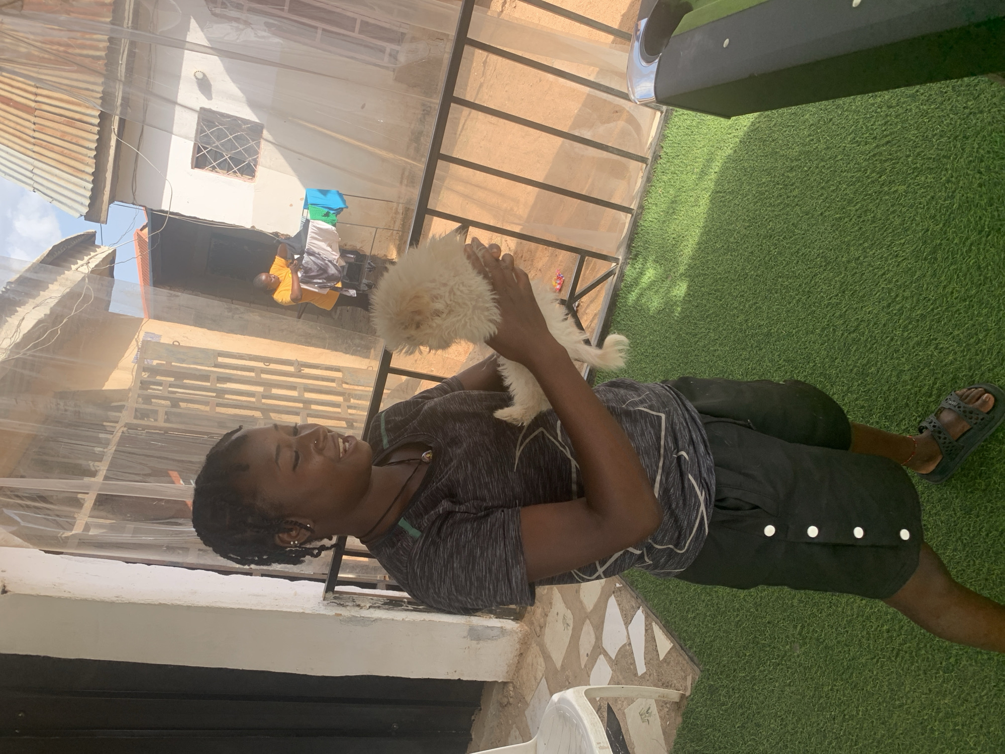 A woman stands outdoors, holding a small, white dog in her hands. She is facing the dog and appears to be smiling. She is wearing a gray t-shirt with a geometric pattern and black shorts with white buttons along the side. She also wears dark sandals.
The setting appears to be a residential area with houses visible in the background, and a corrugated metal roof overhead. There is also green artificial turf underfoot. In the background, another person can be seen wearing a yellow shirt and white shorts. The location is identified as Barakin Akawo, Nigeria.