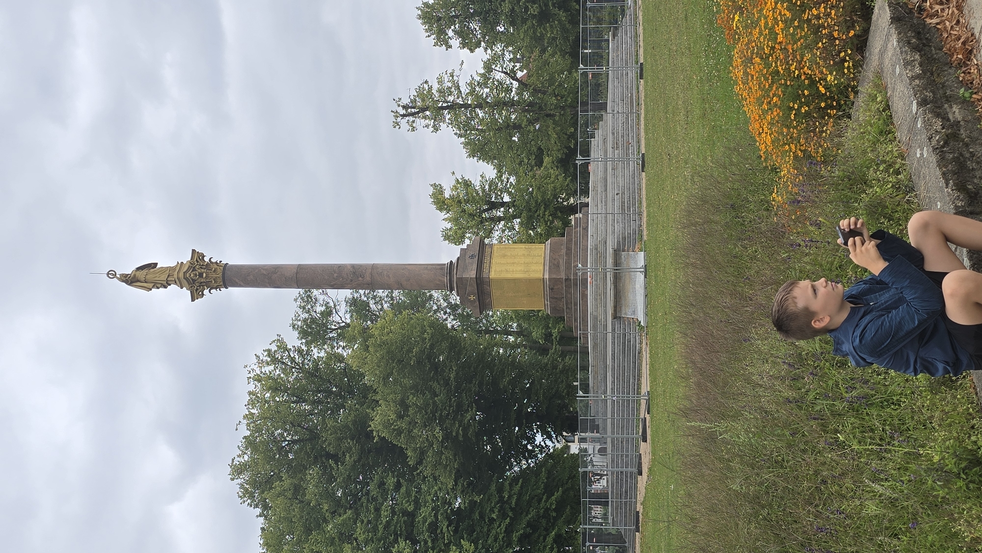 A young boy is sitting on a stone ledge in the foreground, looking at a mobile phone. He is wearing a blue hooded jacket and black shorts. Behind him, a large monument with a golden statue at the top stands in a park. The monument is surrounded by steps and a metal fence. Trees and a cloudy sky complete the scene. The monument is the Victory Column (Siegessäule) located in the Pfaffenteich park in Schwerin, Germany.