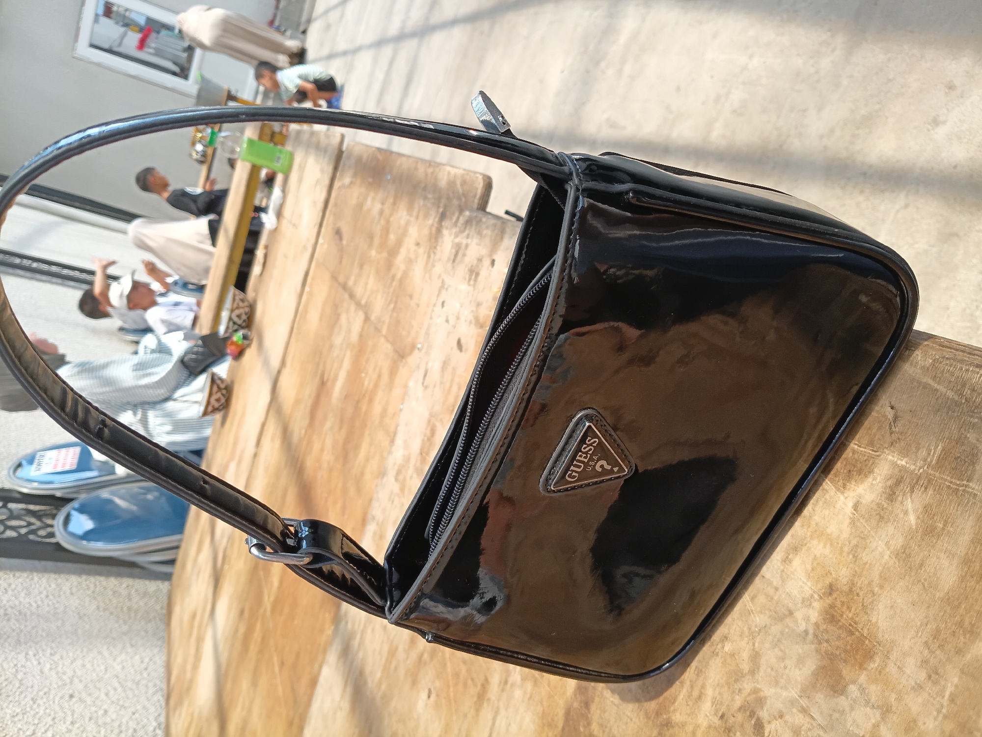 A black, patent leather Guess handbag with a triangular metal logo is placed on a wooden table. The handbag is in the foreground and is the main subject of the image. In the background, several people, including children and adults, are gathered around tables. The setting appears to be an outdoor cafe or market area in Osh, Kyrgyzstan, with some chairs and what looks like a building facade visible. The lighting suggests it is daytime.