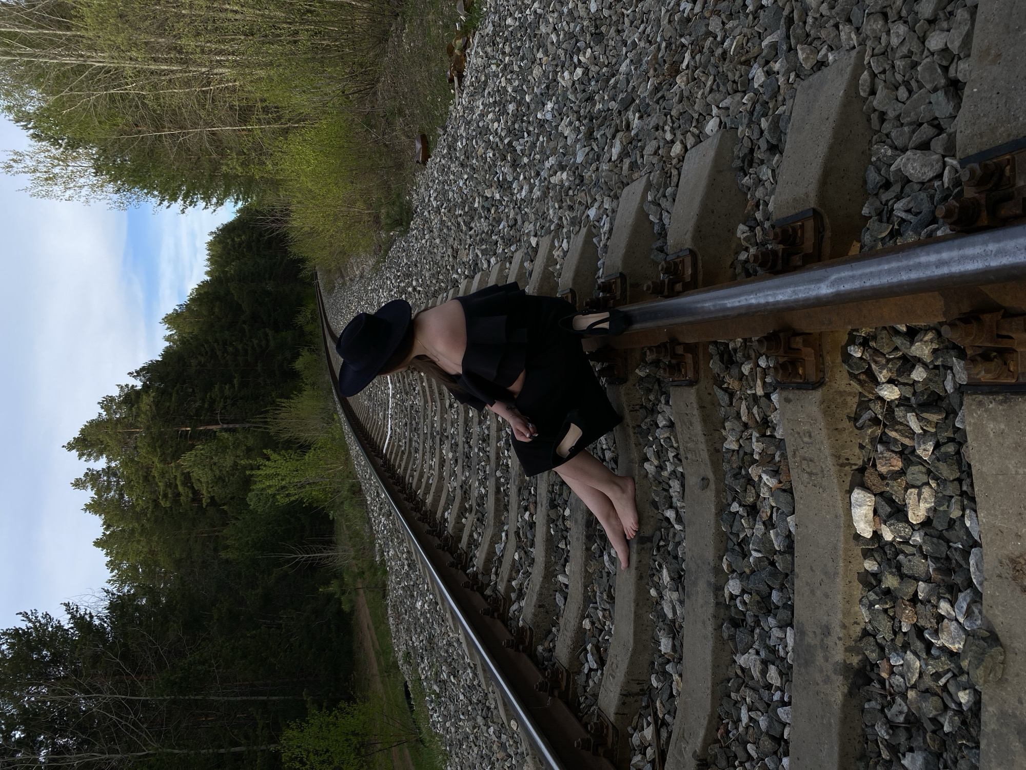 A woman in a black dress and hat poses on a railway track. She is barefoot and leaning on the rail. The tracks are surrounded by gravel and concrete sleepers. Trees and a glimpse of the sky are visible in the background. The specific location in Chaikovsky, Russia cannot be determined from the image.