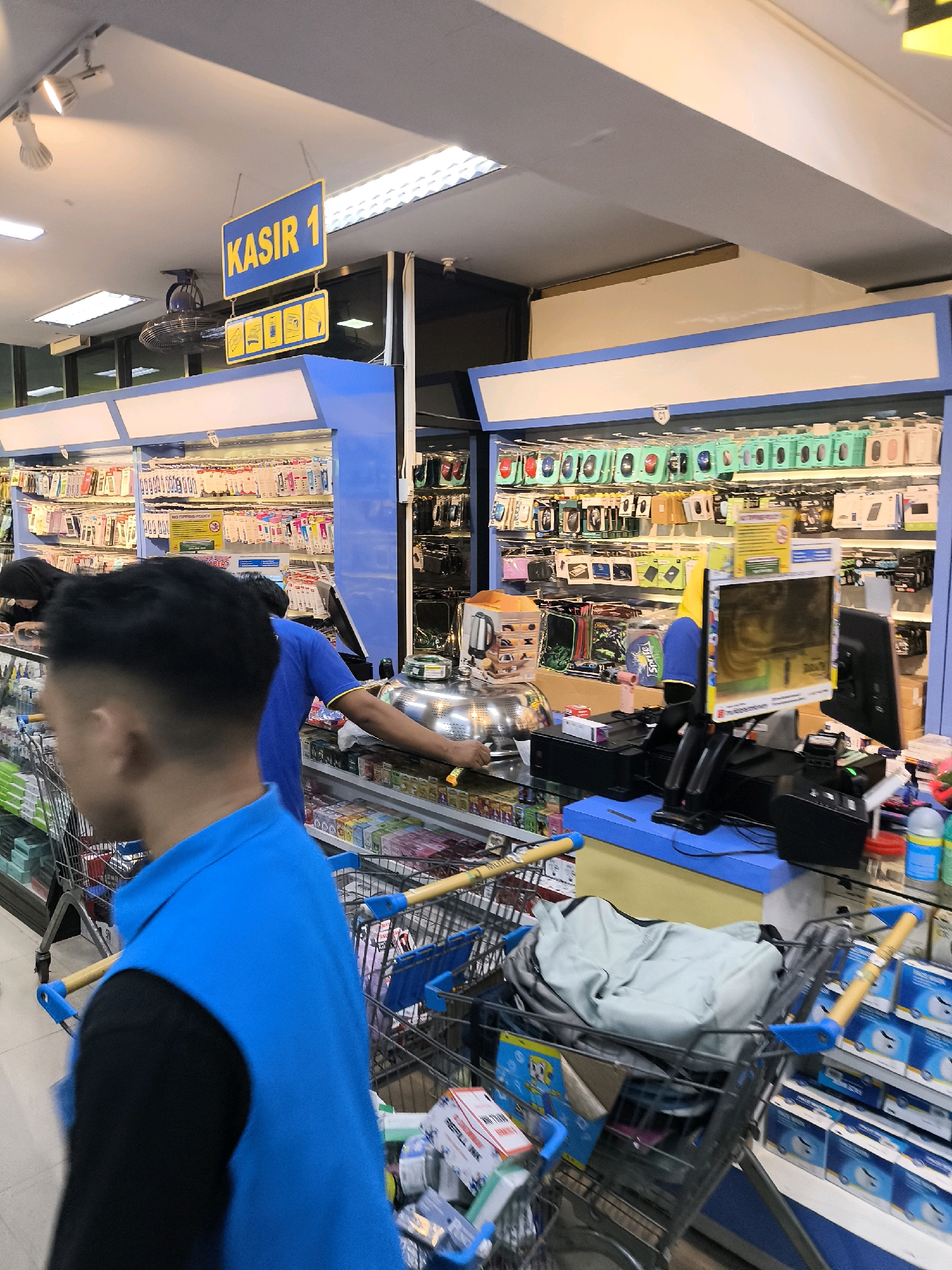The image captures a scene inside what appears to be a retail store, possibly an electronics or general merchandise store, in Gorontalo, Indonesia. A prominent blue and yellow sign hangs from the ceiling, reading "KASIR 1", indicating "Cashier 1". Below this, a smaller yellow sign displays several icons, potentially representing payment methods or product categories.
In the foreground, the back of a person wearing a blue vest over a black shirt is visible, facing away from the camera. This person appears to be pushing a shopping cart filled with items. Another person, wearing a blue shirt, is seen behind a counter, with their arm extended towards a display of goods.
The store environment is characterized by rows of shelves stocked with various products, many of which are small electronic accessories such as phone cases, computer mice, and charging cables, suggesting a tech-focused retail setting. The lighting is bright, likely from overhead fluorescent fixtures. The overall atmosphere suggests a busy commercial space.