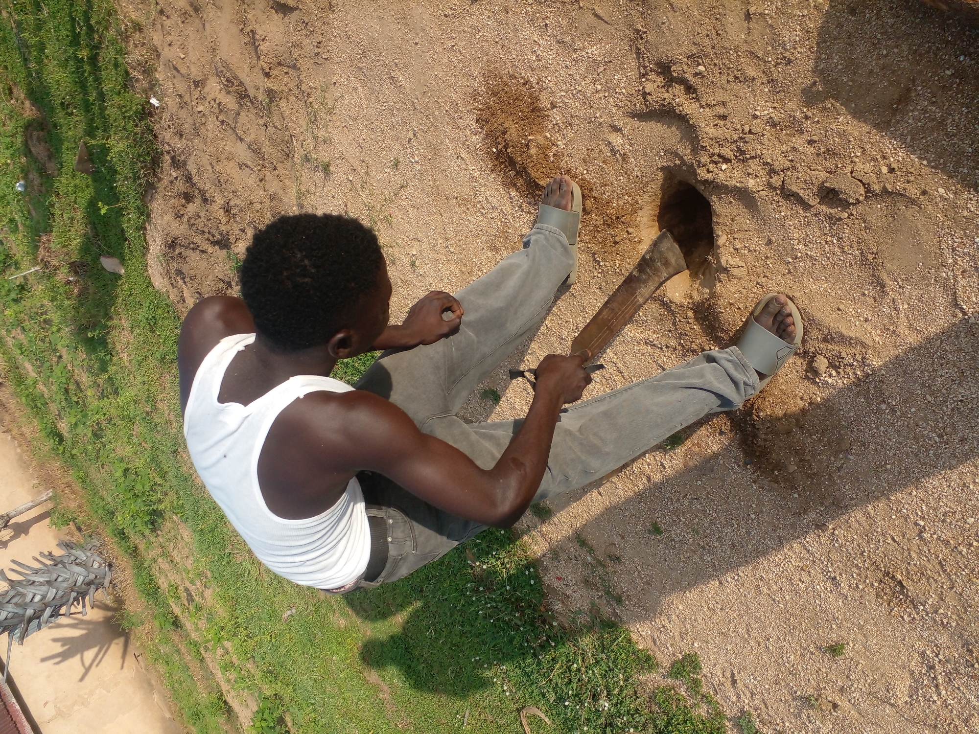 A dark-skinned person wearing a white tank top and grey pants is digging a hole in the sandy ground with a shovel. The person is outdoors in a grassy area with some soil visible. The sun is casting a shadow of the person onto the ground. The location is in the city of Unknown, Unknown.